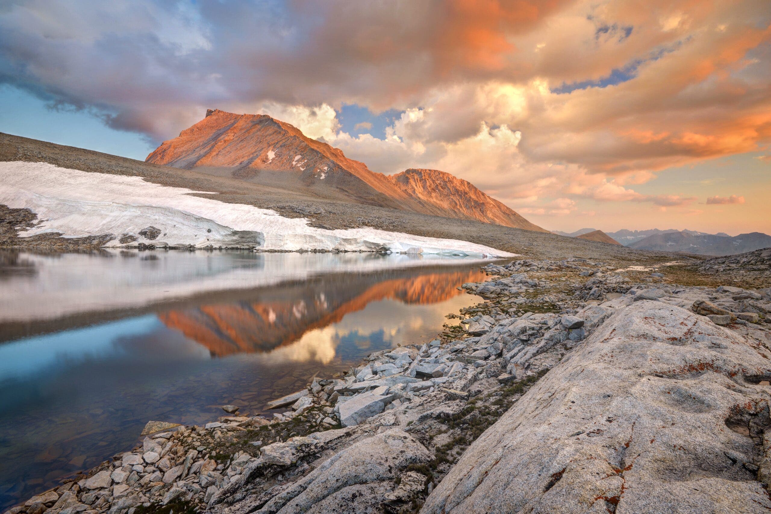 a lake bordered by boulders and snowfields, with a bare rock peak lit up by sunset in the background and reflected in the water