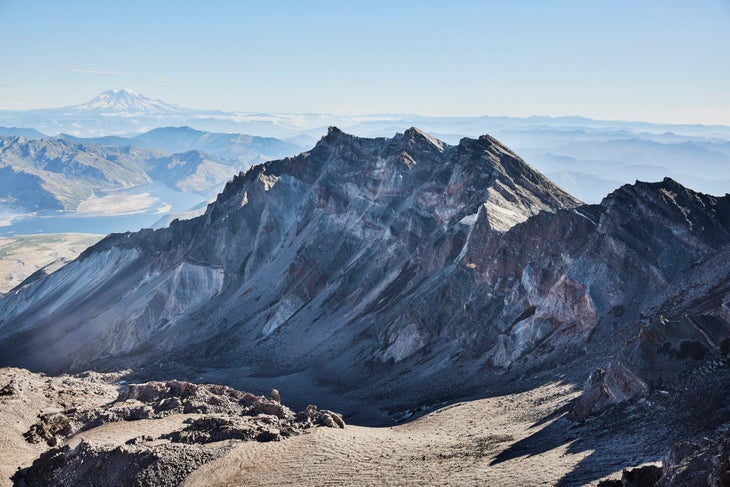 a crater below a jagged gray and reddish rock ridgeline, with the silhouette of Mt Rainier in the far background