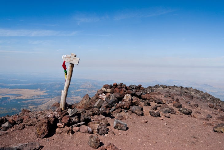 a sandy summit with some rocks and a wooden signpost with some flags hanging off it; in the distnace and below the peak is desert and other mountains