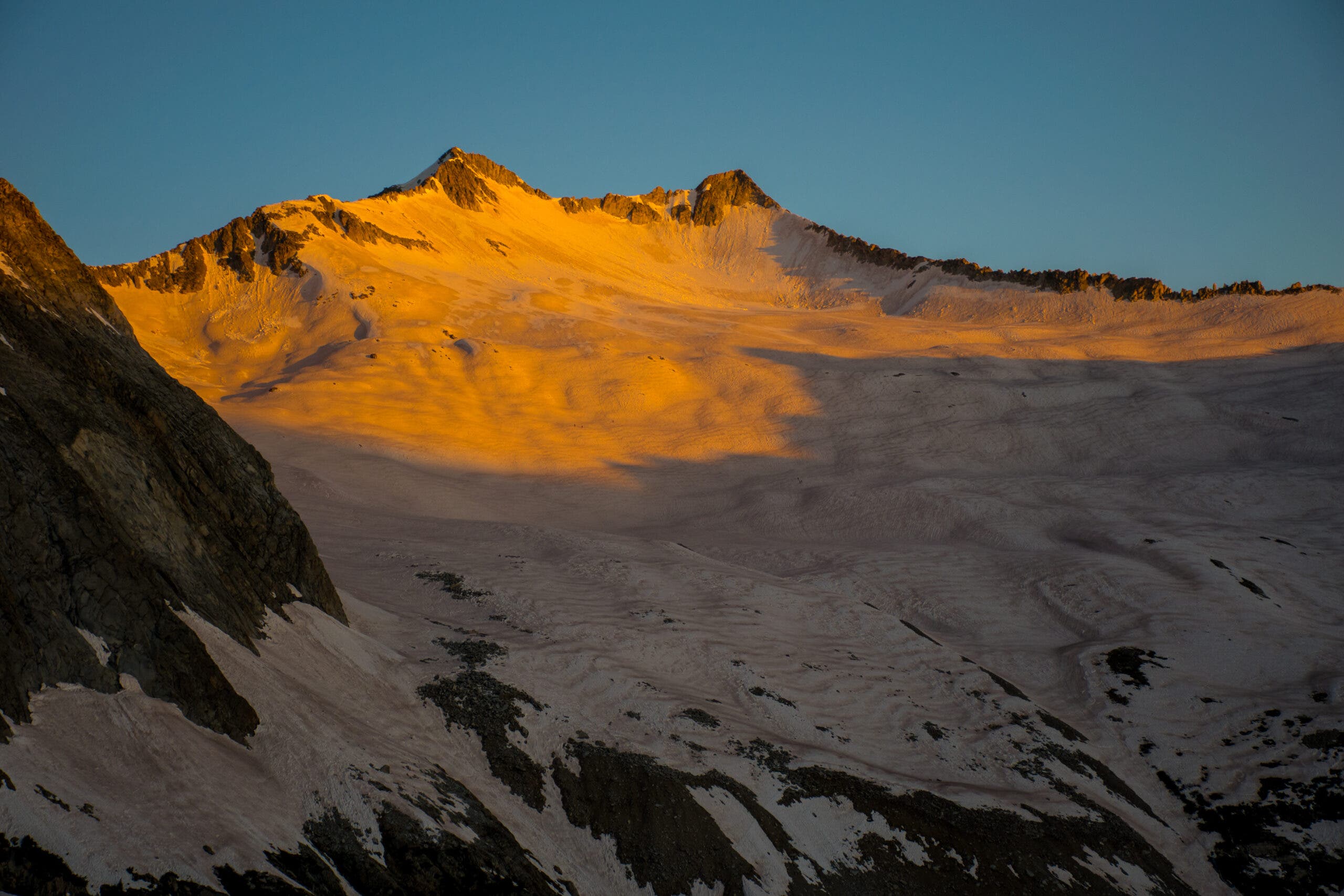 a broad snowfield drops down from a thin ridge with two rocky peaks, the upper half turned gold by early morning light