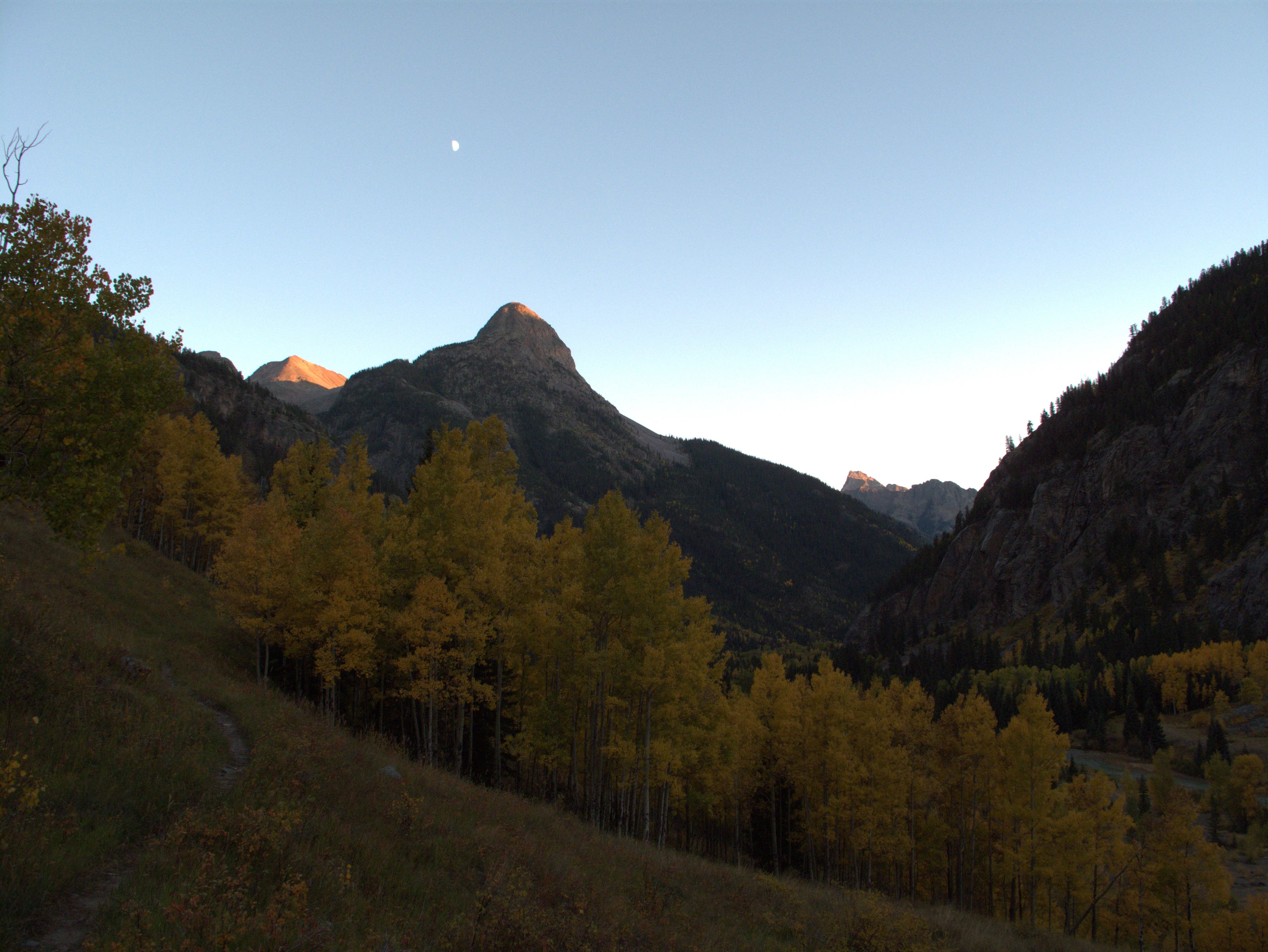 A moon shows itself a dusk above the pointed mountain top of Point Pun. 