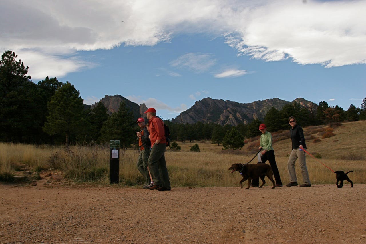 Flatirons views along the Flatirons Loop Trail. Four people walking along the trail with dogs and Flatirons in the distance.