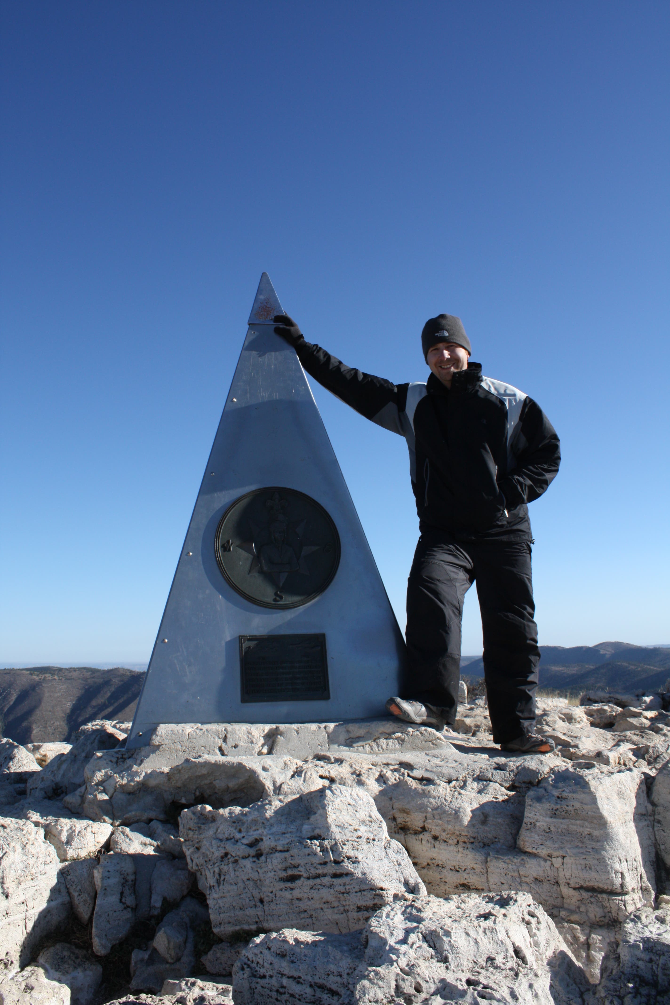 A man standing on the summit of Guadalupe Peak touching the American Airlines Monument, a large silver triangle. 