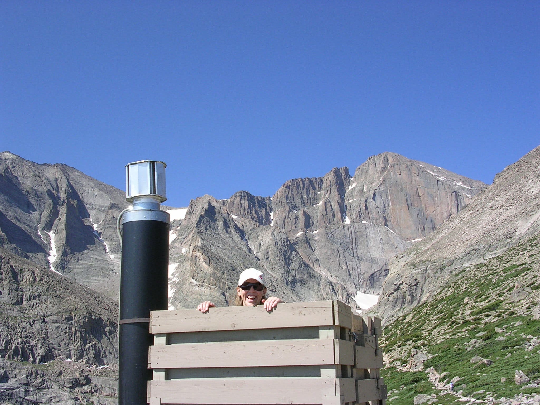 Stop at the Chasm Lake trail privy on your way up. A hiker pops her head out of the top of the privy with Longs Peak in the background.