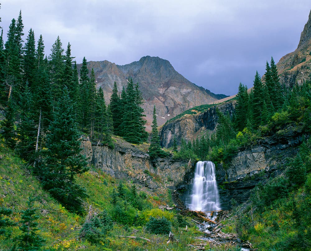 A waterfall cascades over cliffs along Kilpacker Creek in the Lizard Head Wilderness, with El Diente Peak (14,159’) rising in the distance.