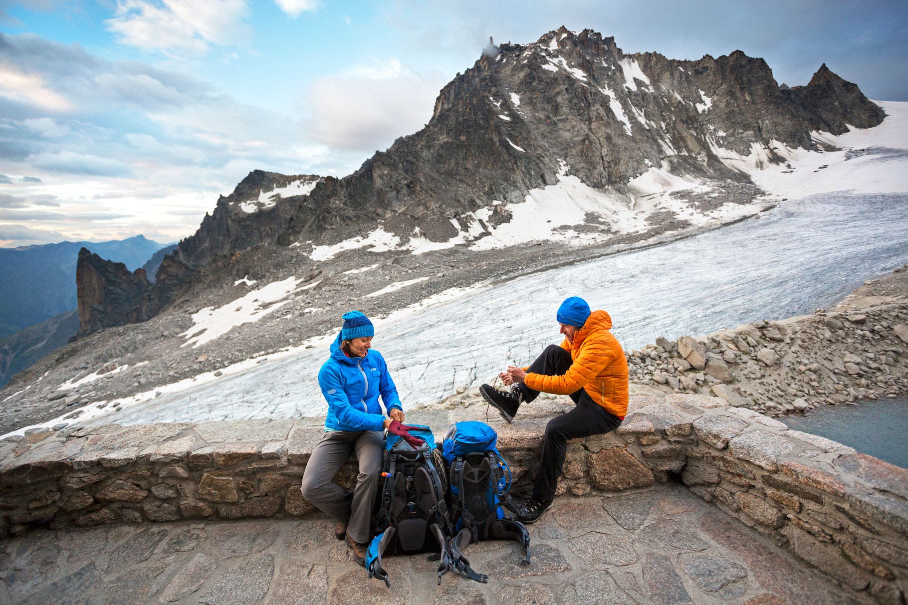 Two hikers sitting on the deck of the Cabane d'Orny tying shoes and getting ready to leave for the second day of the Chamonix to Zermatt Glacier…