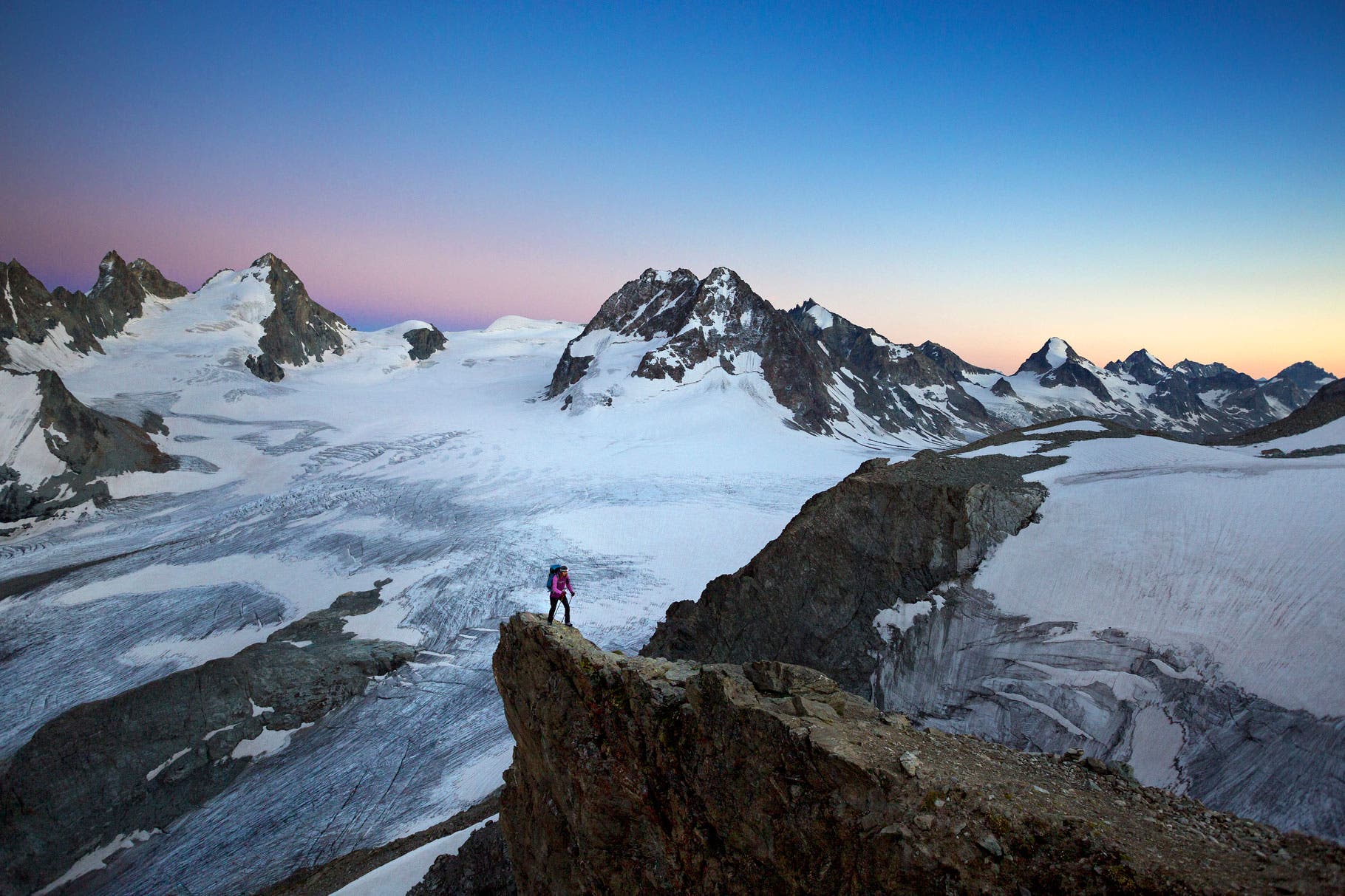 A hiker at sunset high above glacier covered mountains using a headlamp while walking to the Vignette Hut on the third day of the Chamonix to Zermatt…