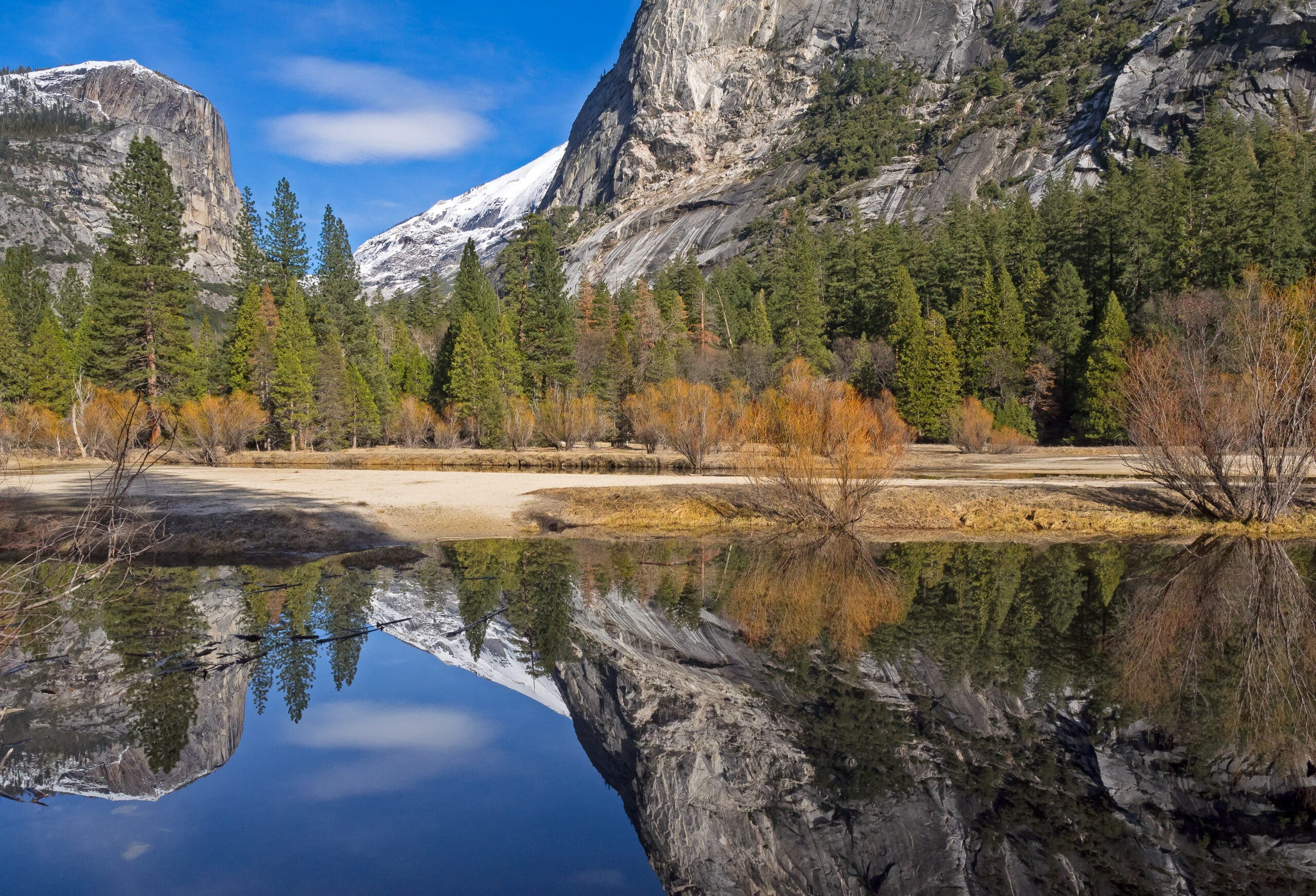 a calm lake in the foreground reflects evergreen trees in front of a steep granite cliff, with snowy hills behind