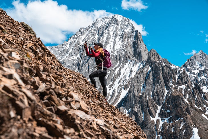 A female hiker ascends a steep scree slope with trekking poles in front of a mountain.