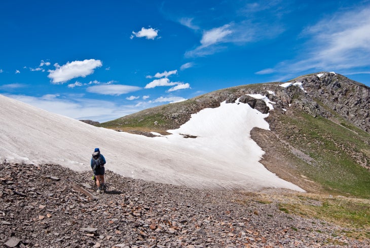 Hiker standing beside a snowfield on scree atop a mountain pass.