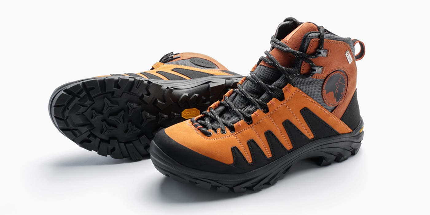 Orange and black Mishmi hiking boots sit on a white background. One is upright on its sole and the right shoe is lying on its side with the Vibram…