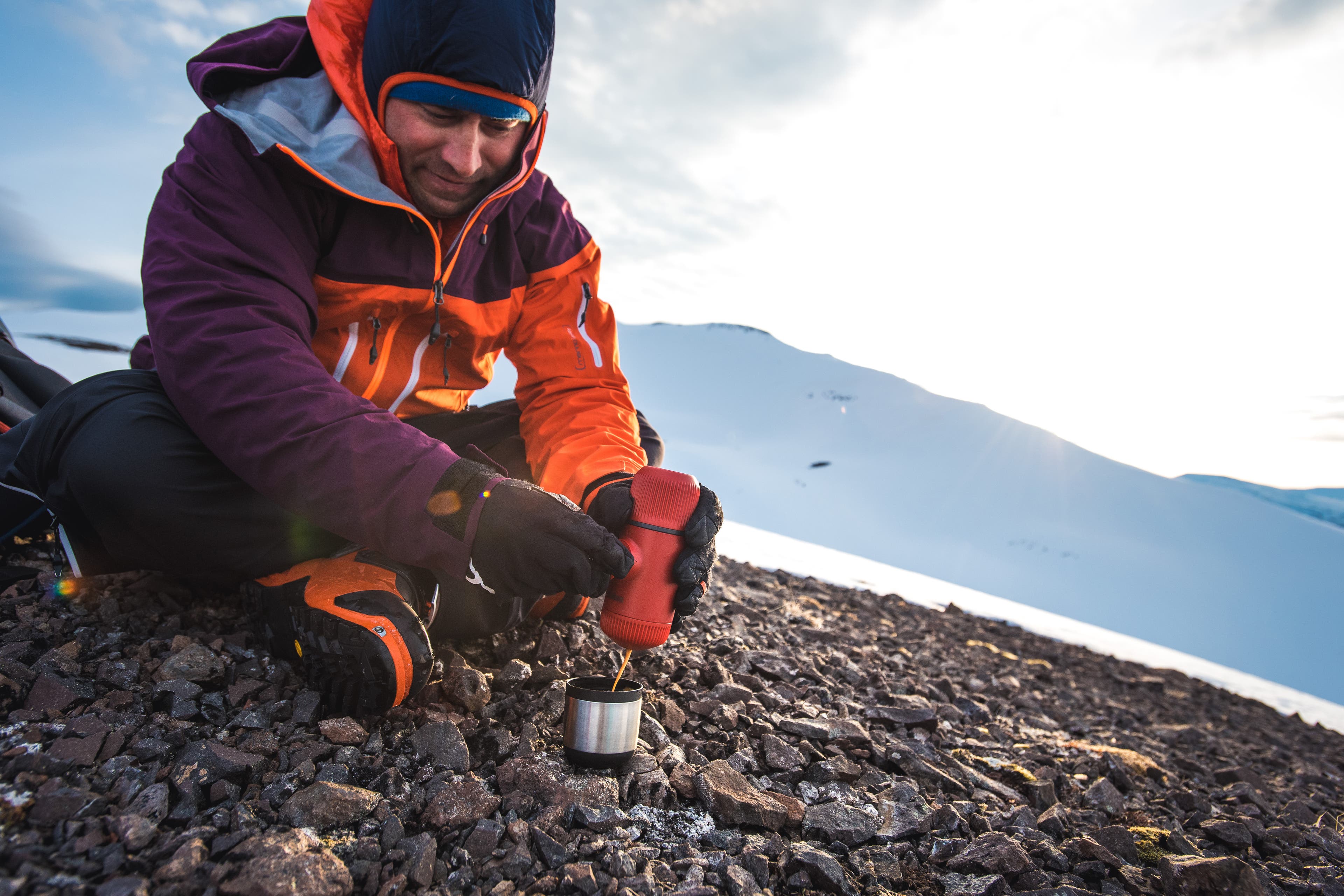 Nanopresso_coldweather Man sits on rocky ground bundled in orange, black, and purple clothes and makes a cup of espresso with Nanopresso. Mountains peak up in the…