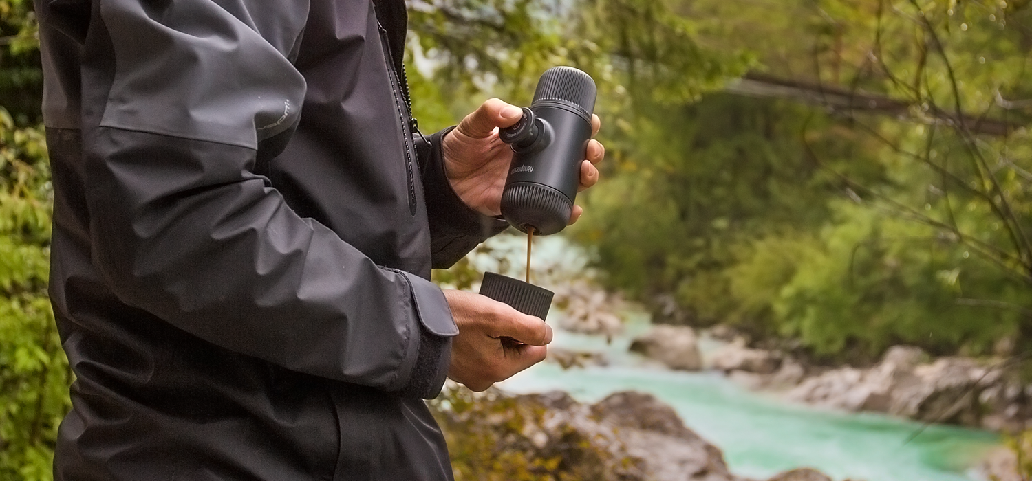 "Person in black jacket standing by turquoise river surrounded by deciduous trees makes a cup of espresso with Wacaco's Nanopresso."