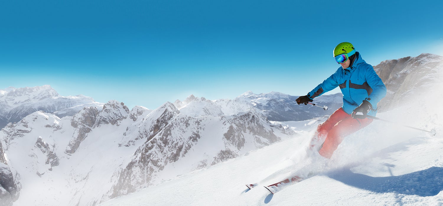 "Man in bright blue jacket, lime green helmet, and orange pants wearing a Casio Pro Trek watch skis atop a powdery mountain. Jagged snowy mountains&hellip;"