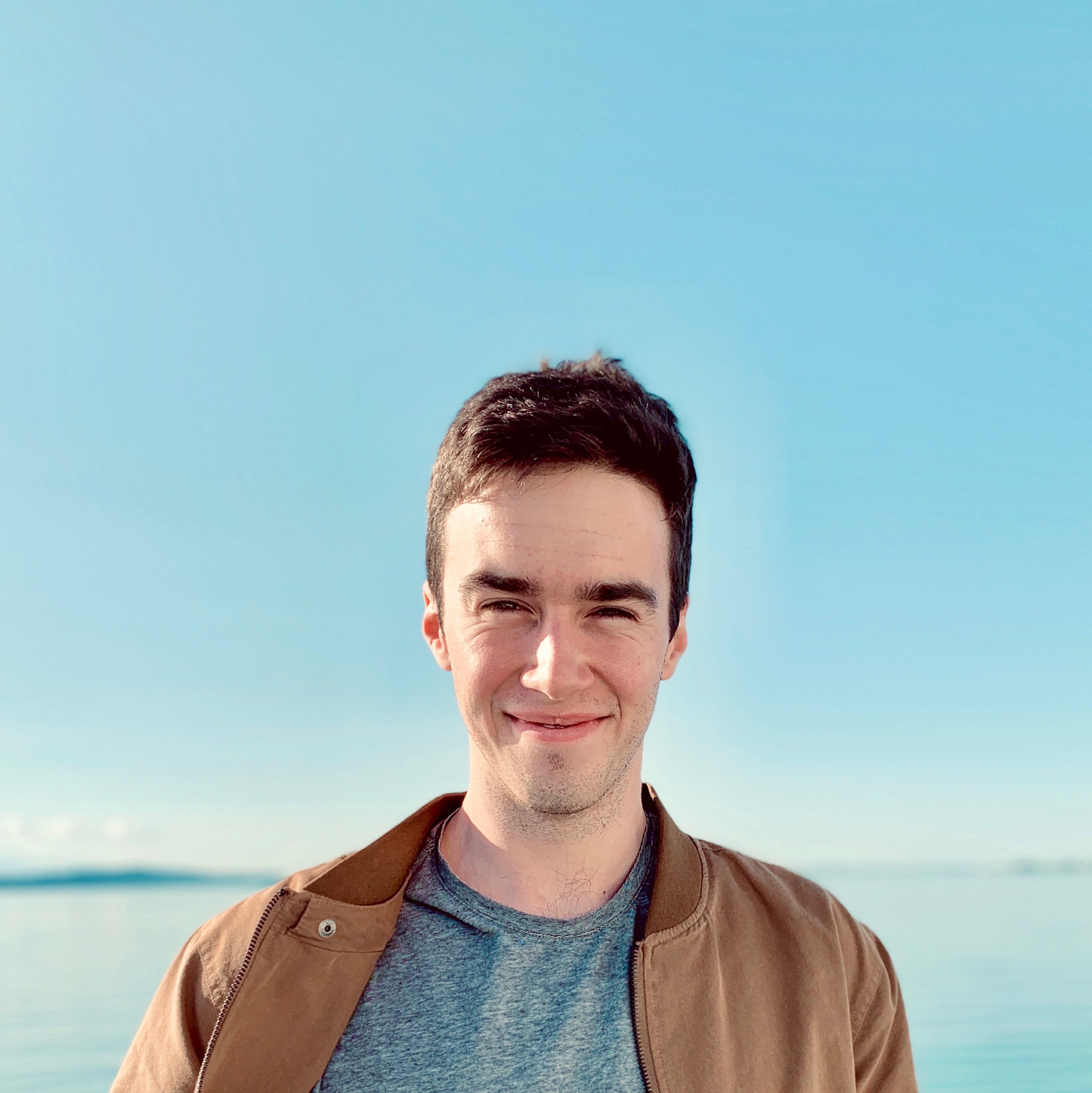 Dave Bonan smiles at the camera in a brown leather jacket with a clear blue sky and body of water in the background.