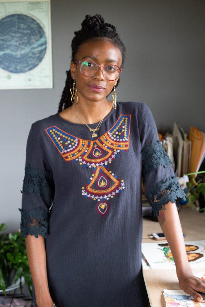 Latasha Dunston stands near her art in a muted navy blue tunic shirt with a geometric red, orange, blue and white design on the front. She wears gold…