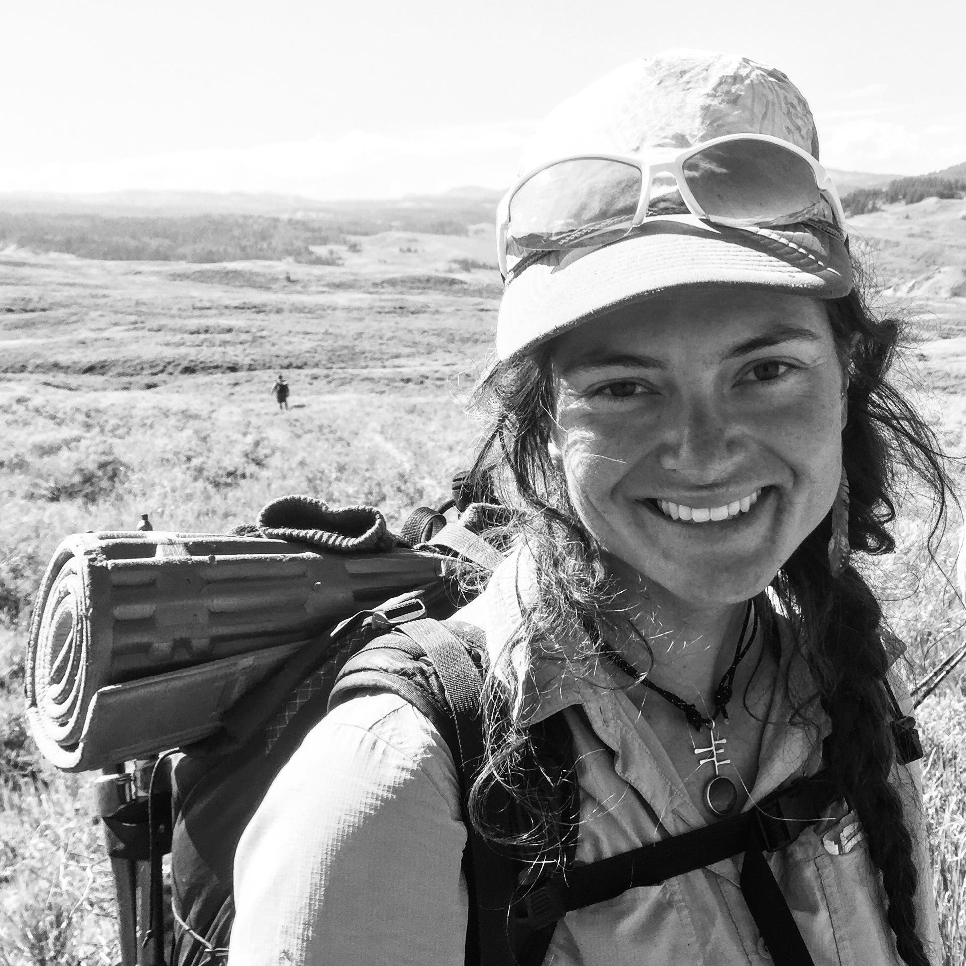 Nika Meyers smiles at the camera in a black and white photo with a grassy background. She is wearing a hat with sunglasses on the brim, and a…