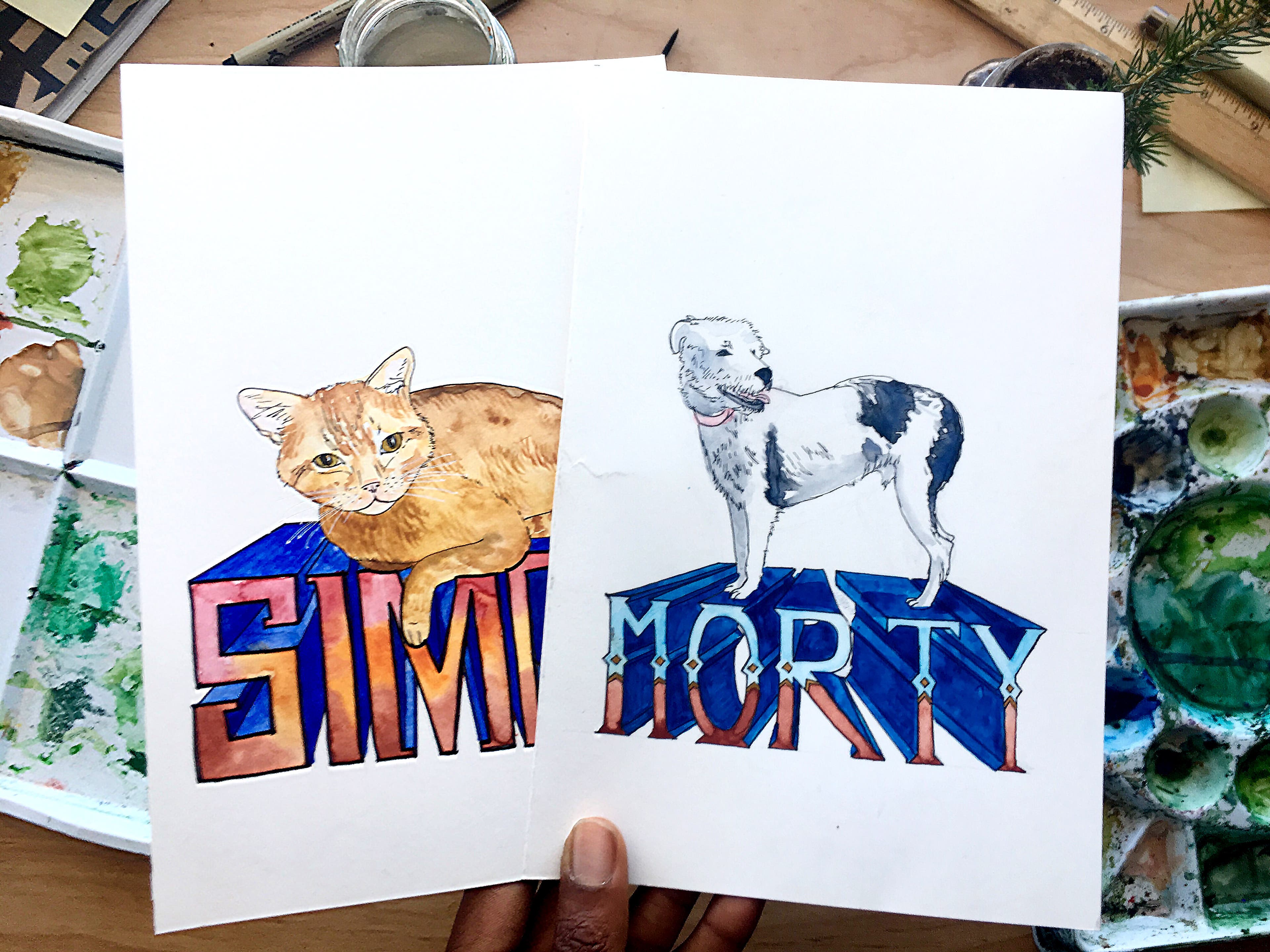 Two pet portraits sit in a hand over a background of paints on a table. One is of an orange cat and the other is a dog. Both pets stand on 3D letters…