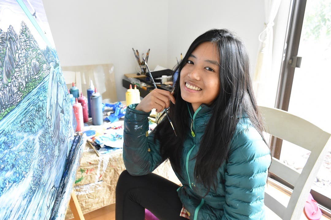 AlexRubio Alex Rubio, an asian american woman, sits in a white chair smiling with a paint brush in her hand facing a canvas with a partially painted landscape.
