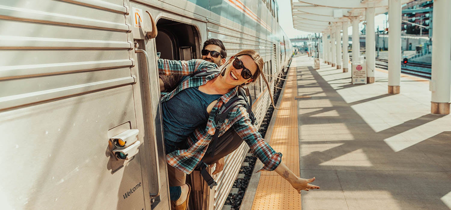 "White woman in plaid long button down with a Gregory Proxy pack on her back hangs out of a train window."