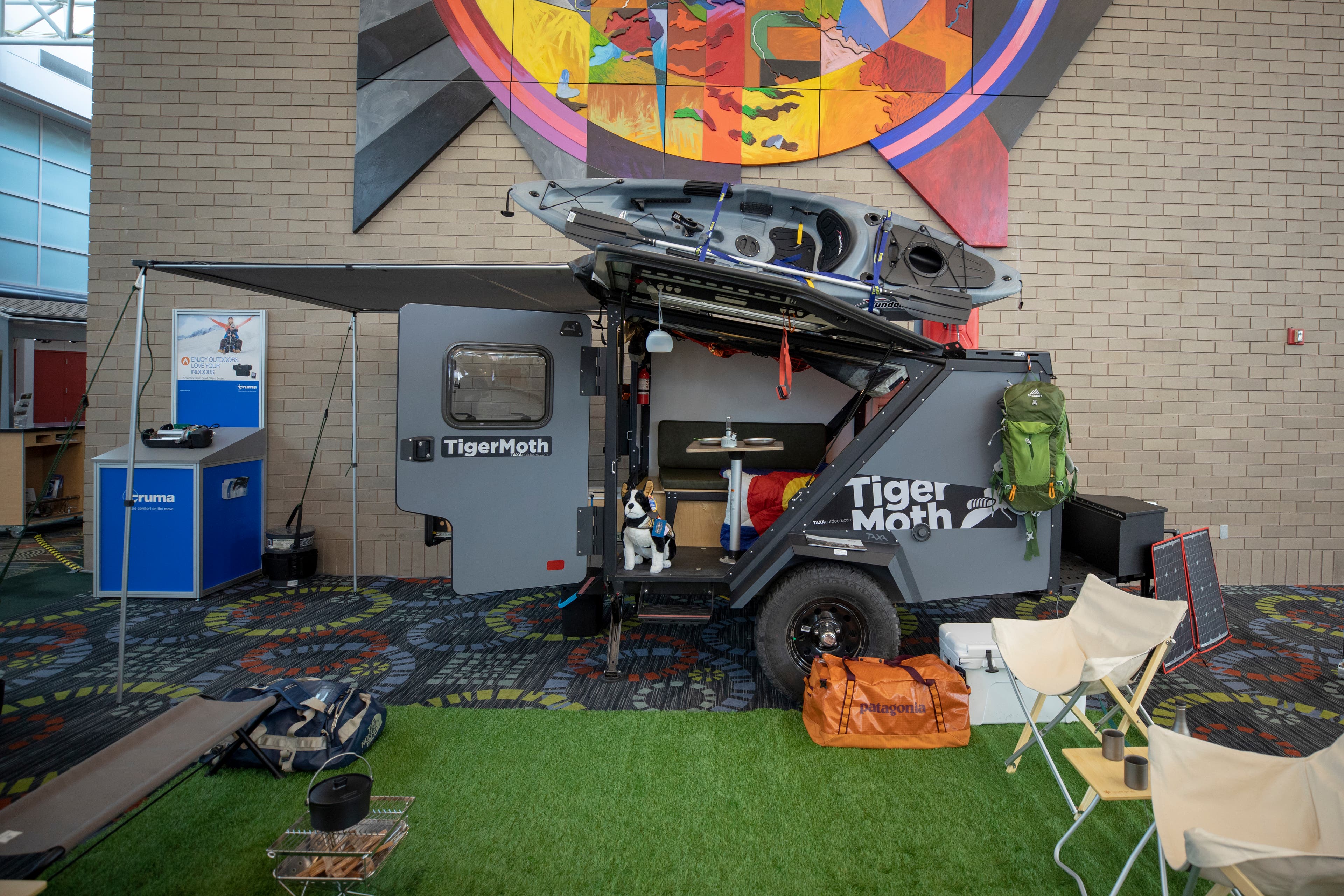 Taxa TigerMoth RV sits on the RVX show floor with doors open, a kayak on top, and camp chairs set up outside.