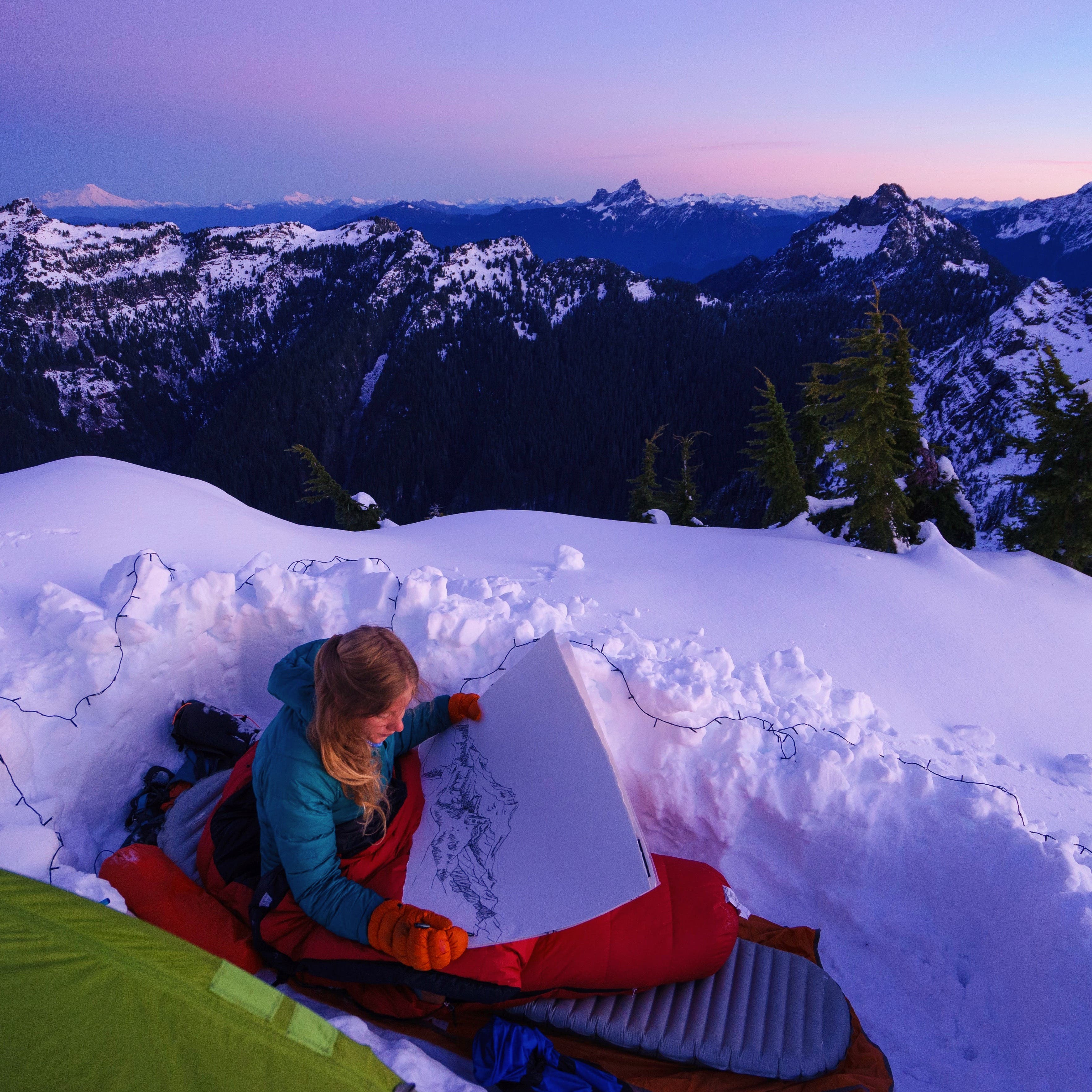 White woman sits in a sleeping bag on a mountain top while drawing.