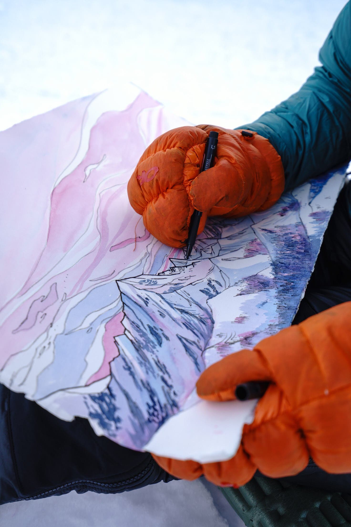 Gloved hands hold a pen while drawing a mountain scape.