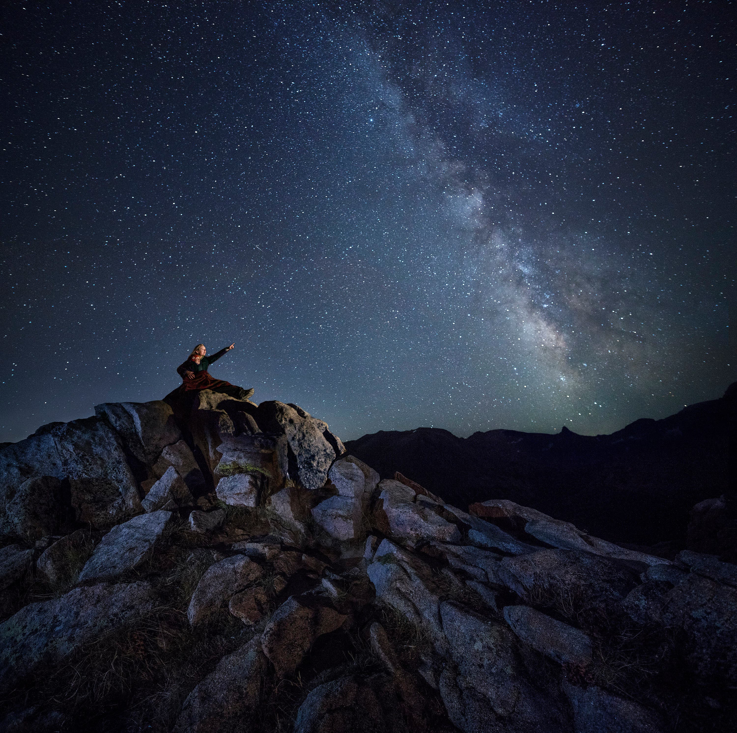 A parent and child sit on a rock outcropping and point at a clear night of stars above them.