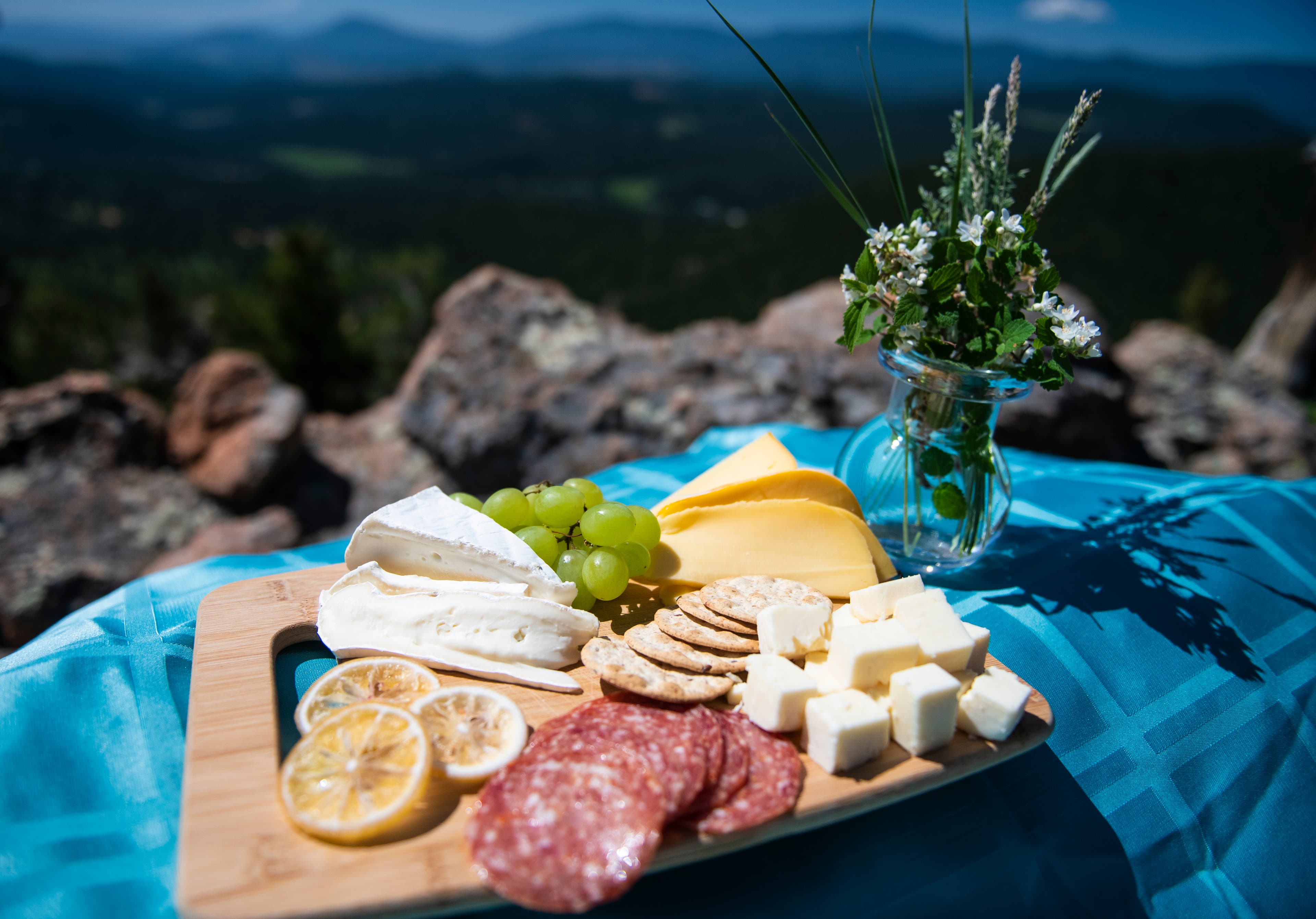 Charcuterie board of salami, brie cheese, crackers, gouda, and cheddar cubes sit on a teal table clothe with a flower vase next to it. Rocks and…