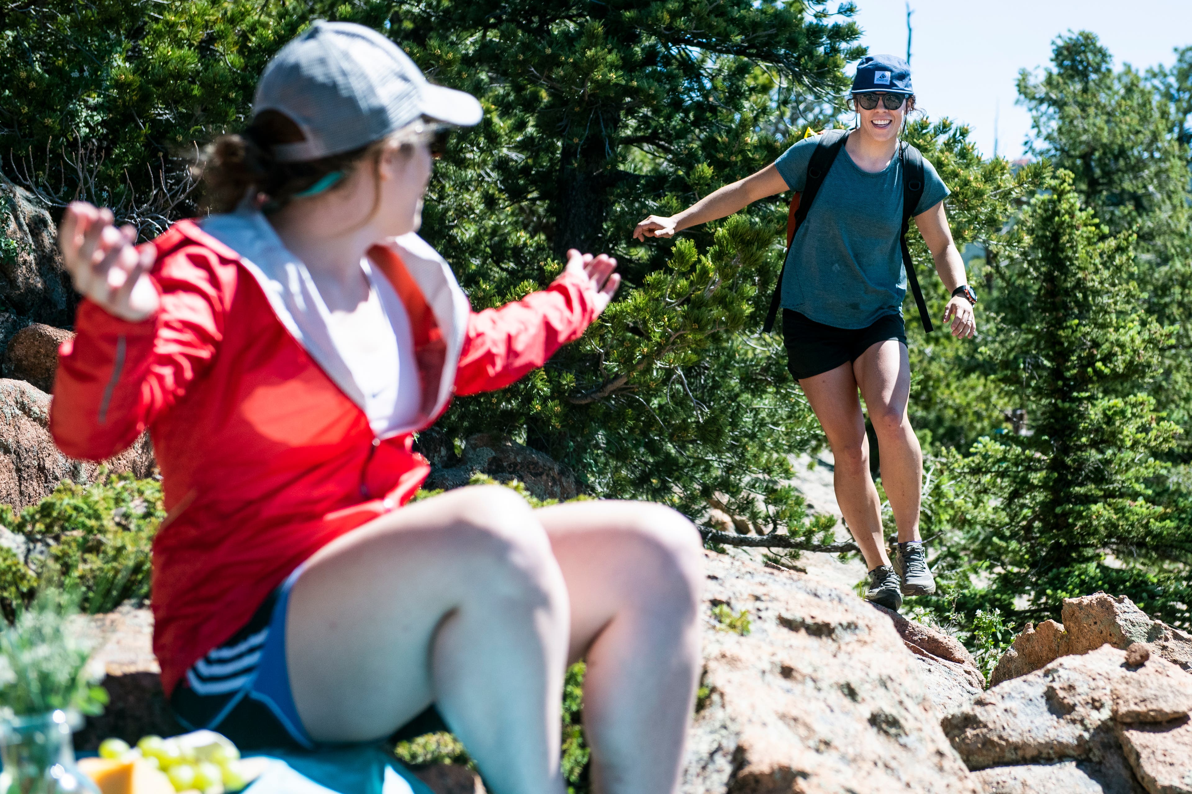 White woman in muted blue shirt walks along edge of rocks toward another white woman in a red jacket sitting down on the rocks.