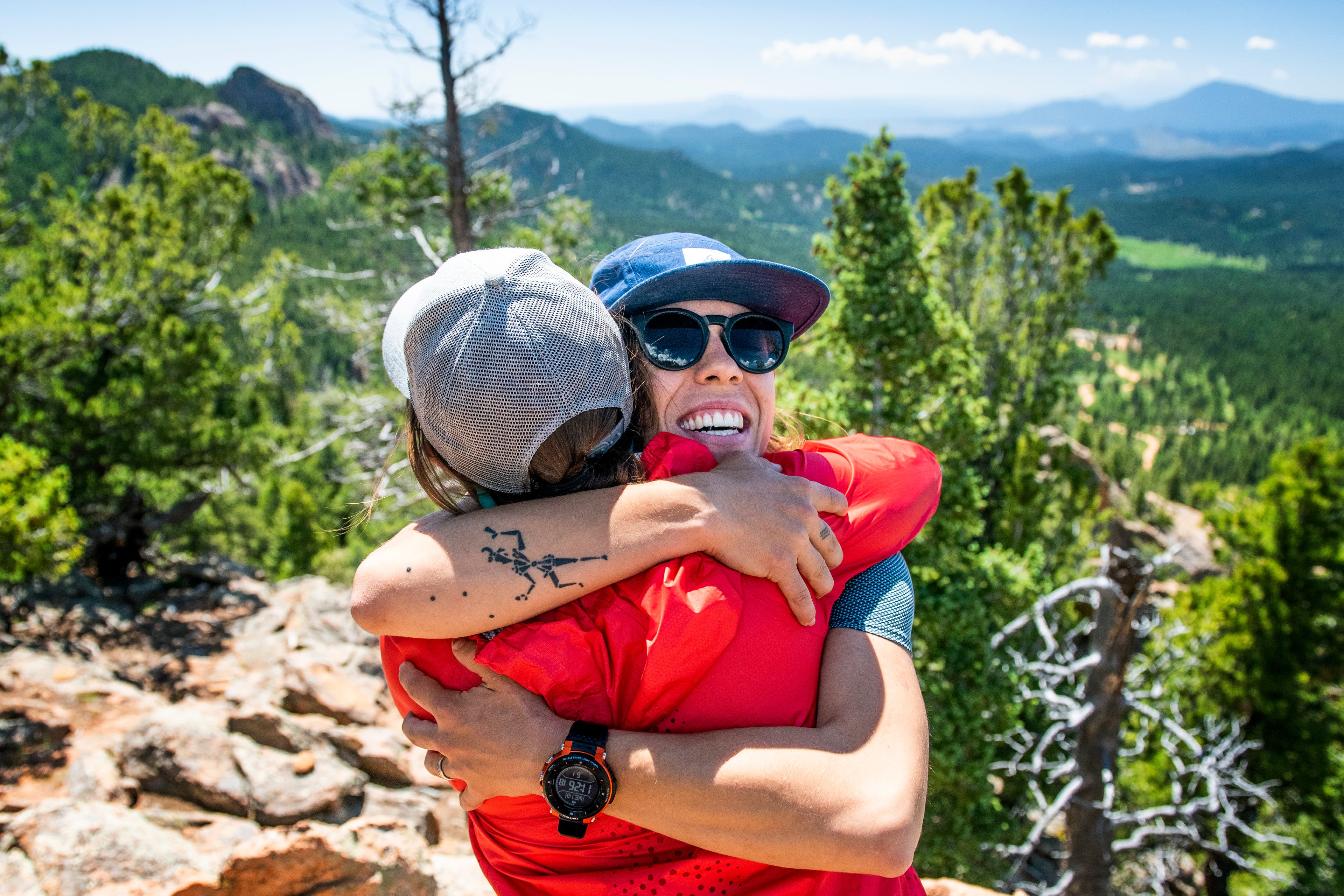 20190710_ViewRanger_568 Two women hug. One in a blue hat and smiling toward the camera, the other in a red jacket and gray hat facing the mountains.