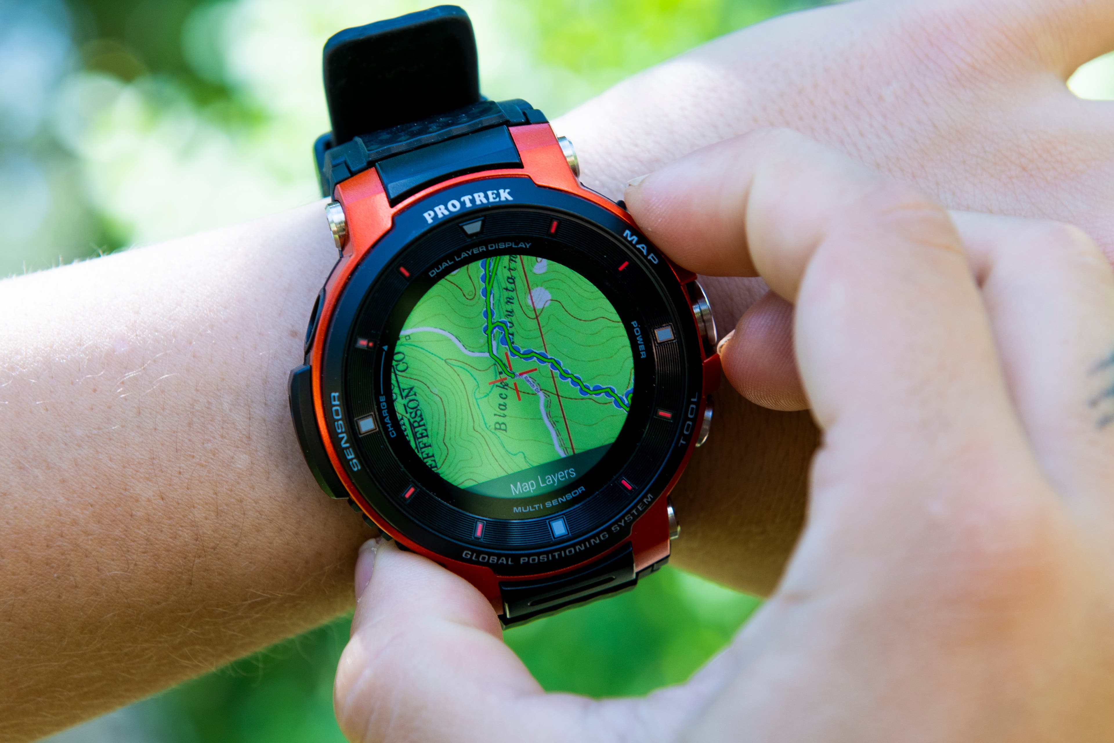 Close up shot of white hands adjusting the ViewRanger app on a black and orange Casio ProTrek watch. A green topographic map with a blue route…
