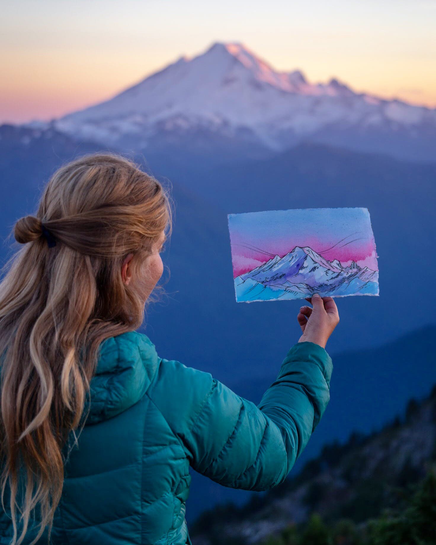 Blonde woman holds up painting of Mount Baker with the mountain and sunset in the background.