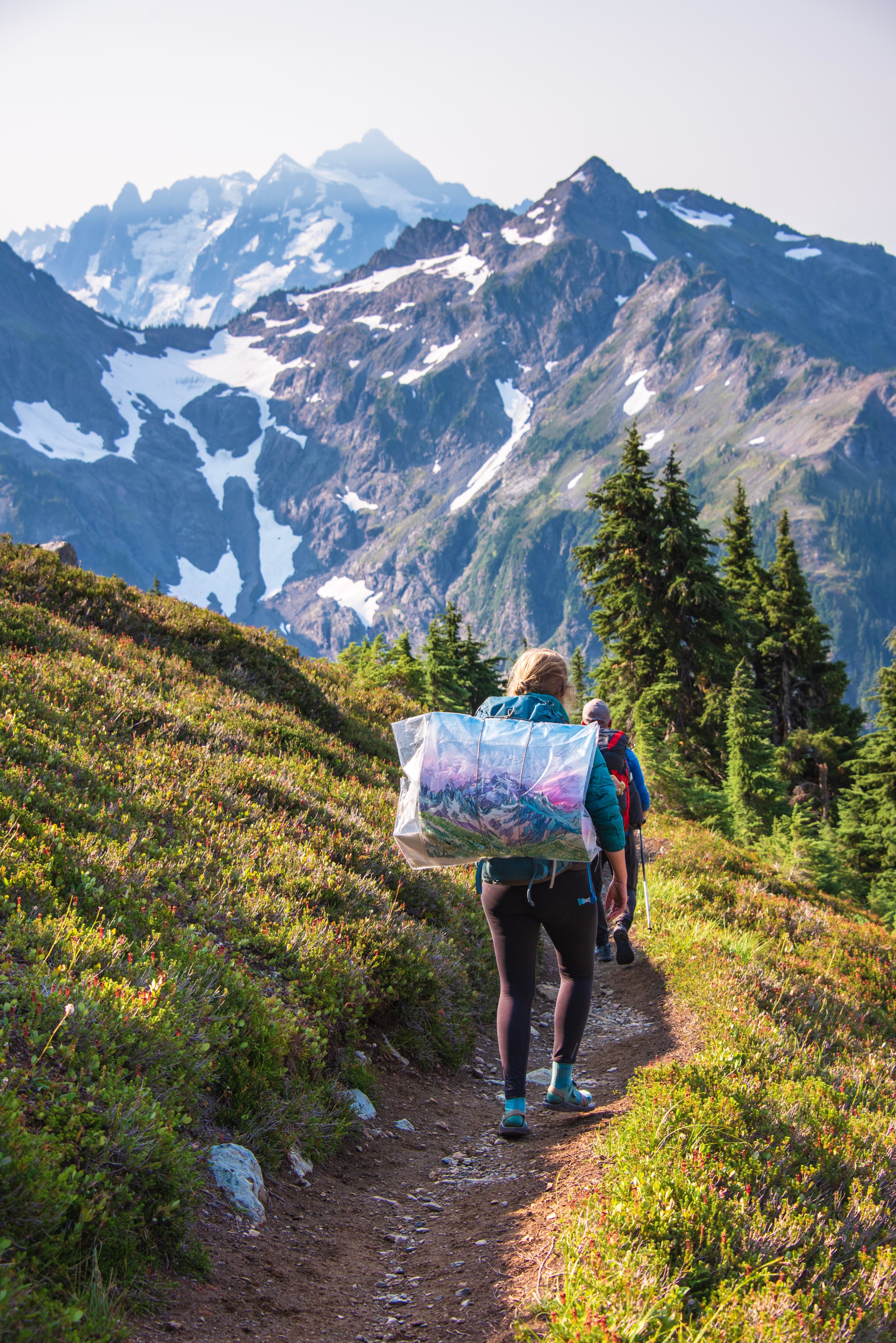 Woman hikes with mountain views and painting strapped to her backpack.