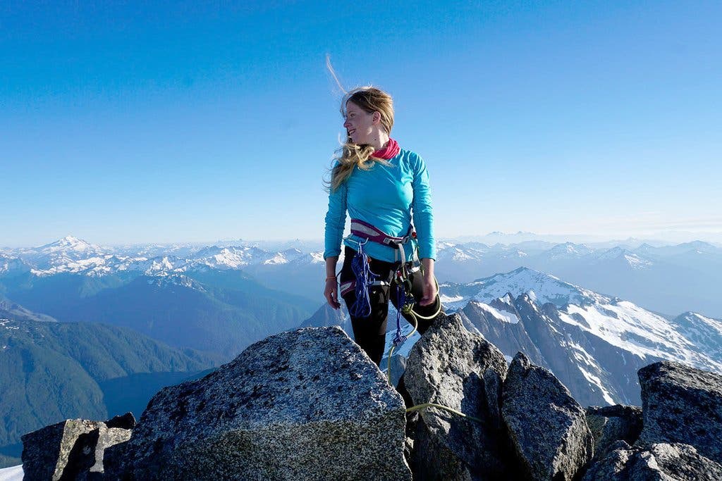 Nikki Frumkin, a blonde white woman, stands on top of a mountain in a climbing harness with her hair blowing in the wind. Blue skys and snow-capped…