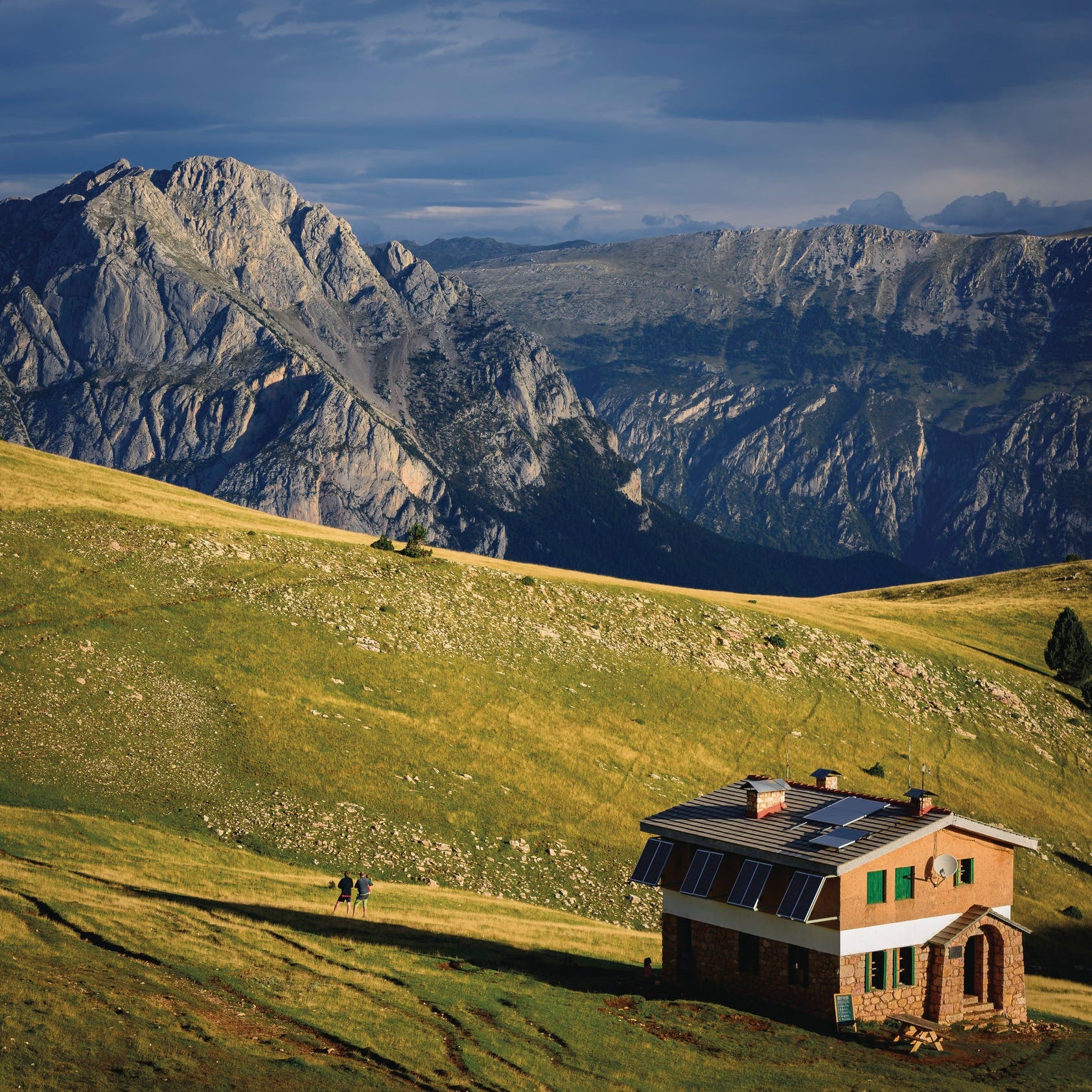 Cavalls del Vent-Ensija Shelter @CTB_Sergi Boixader Bright yellow-green grass covers hills in the foreground where two people stand near a small brick shelter and look out on gray rocky mountains in…