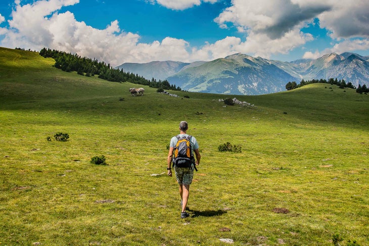 @CTB_INMEDIA SOLUTIONS White man with a bakcpack and cargo shorts walks through a grassy field toward a cliff overlooking mountains. La Porta del Cel in Catalonia, Spain