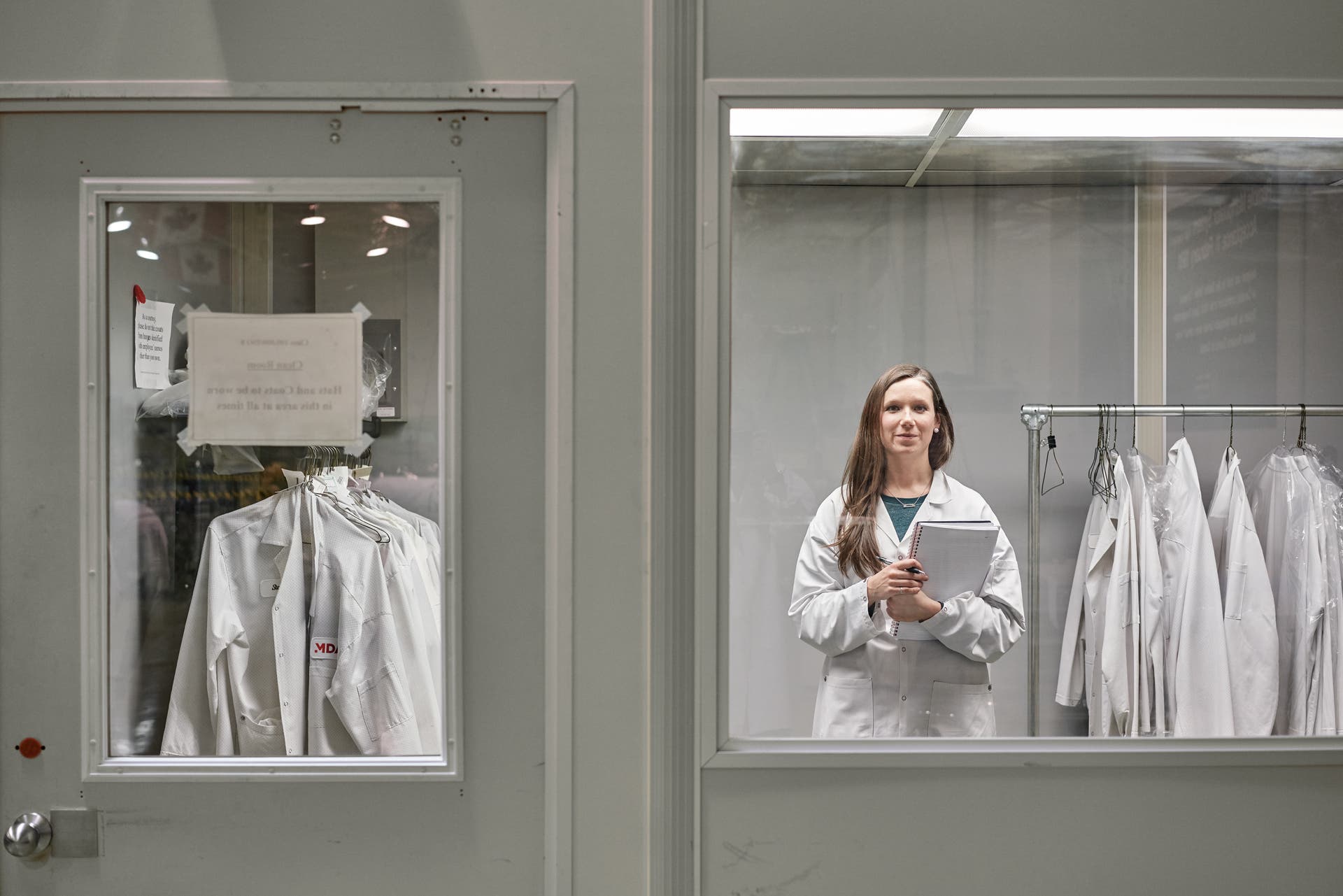 190201_NatPanekToronto_05226shrp White woman in a lab coat facing the camera through a window holding a notebook.