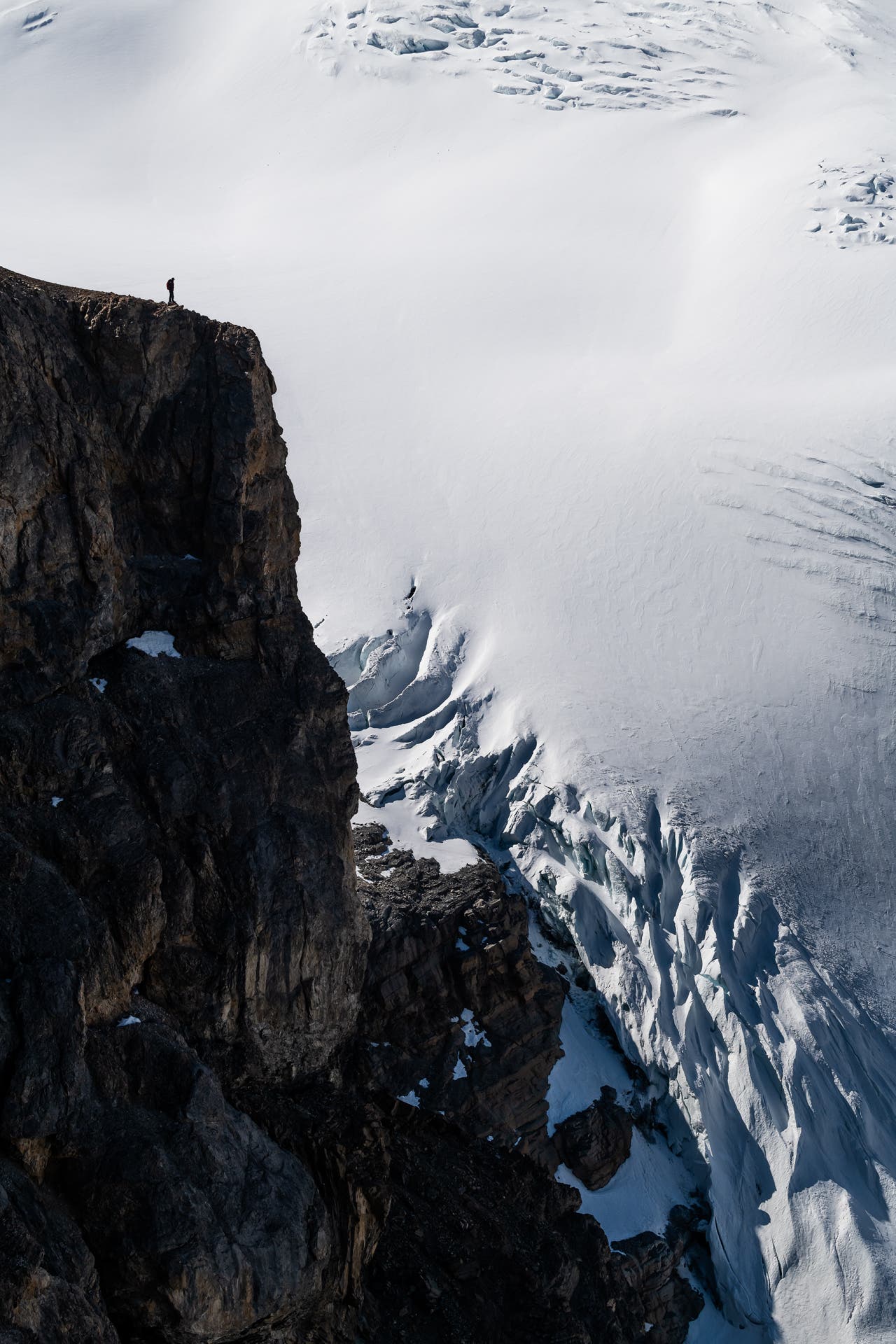 Portrait oriented photo of a rocky cliff with a snowy mountain wall behind it. A small silhouette of a woman stands near the edge.