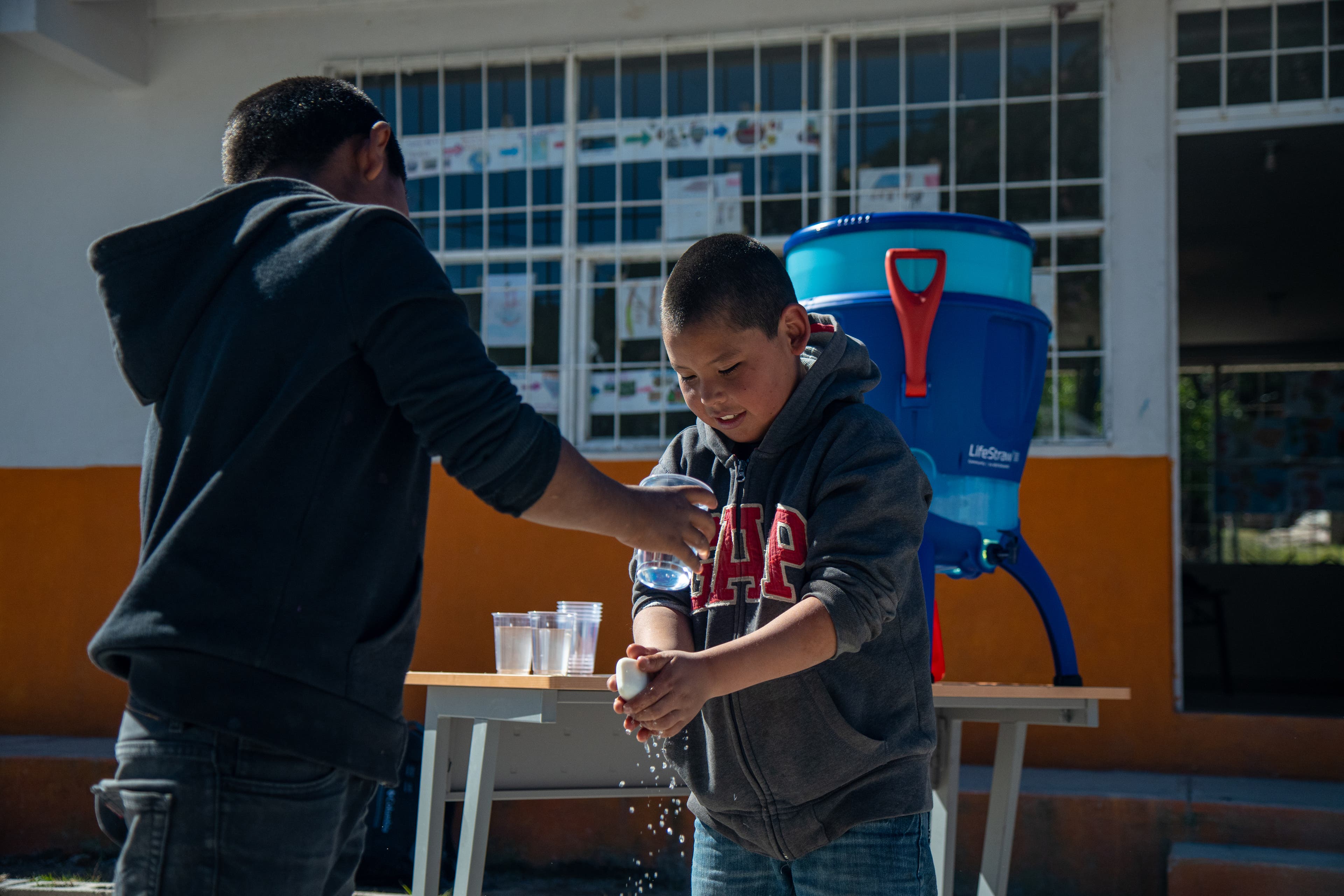Two Tarahumara Indian boys in GAP sweatshirts wash their hands with the clean water in the courtyard outside of school.