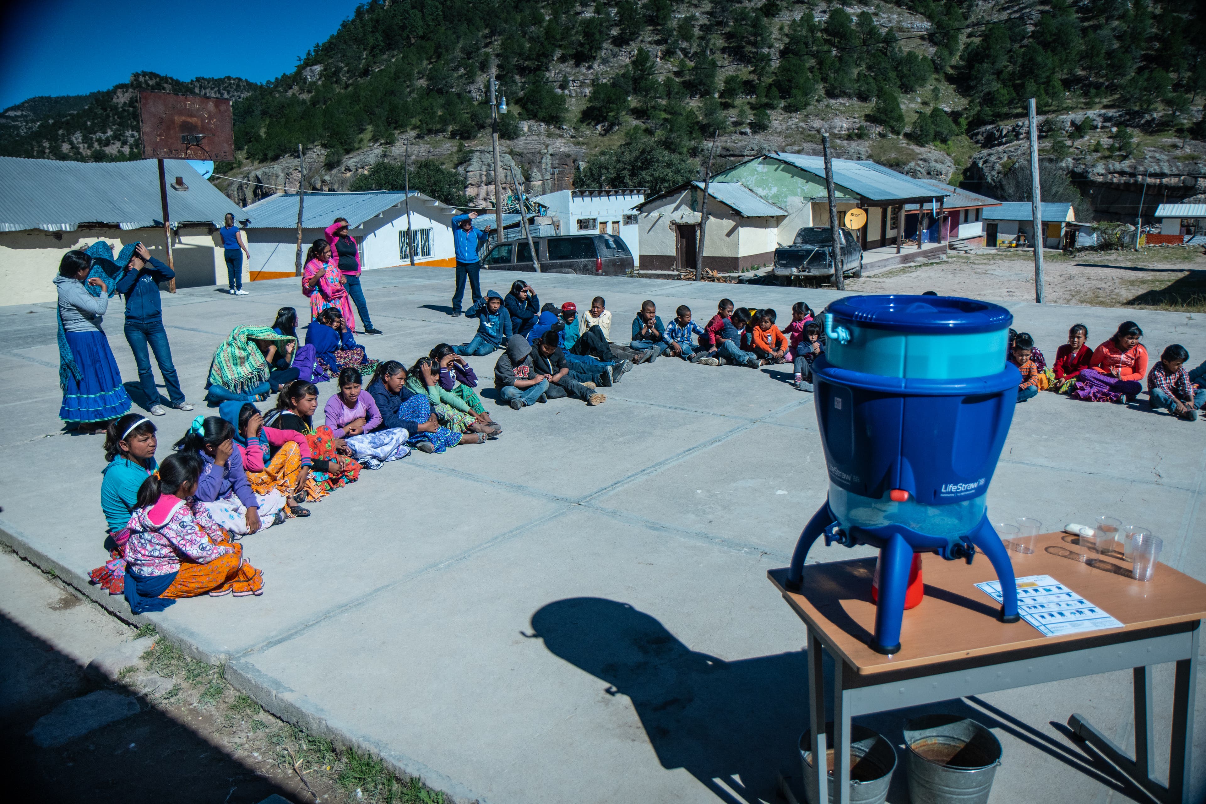 Lifestraw water filter cooler sits on a desk on a concrete area where Tarahumara kids sit awaiting a presentation of the filter. Houses and mountains…