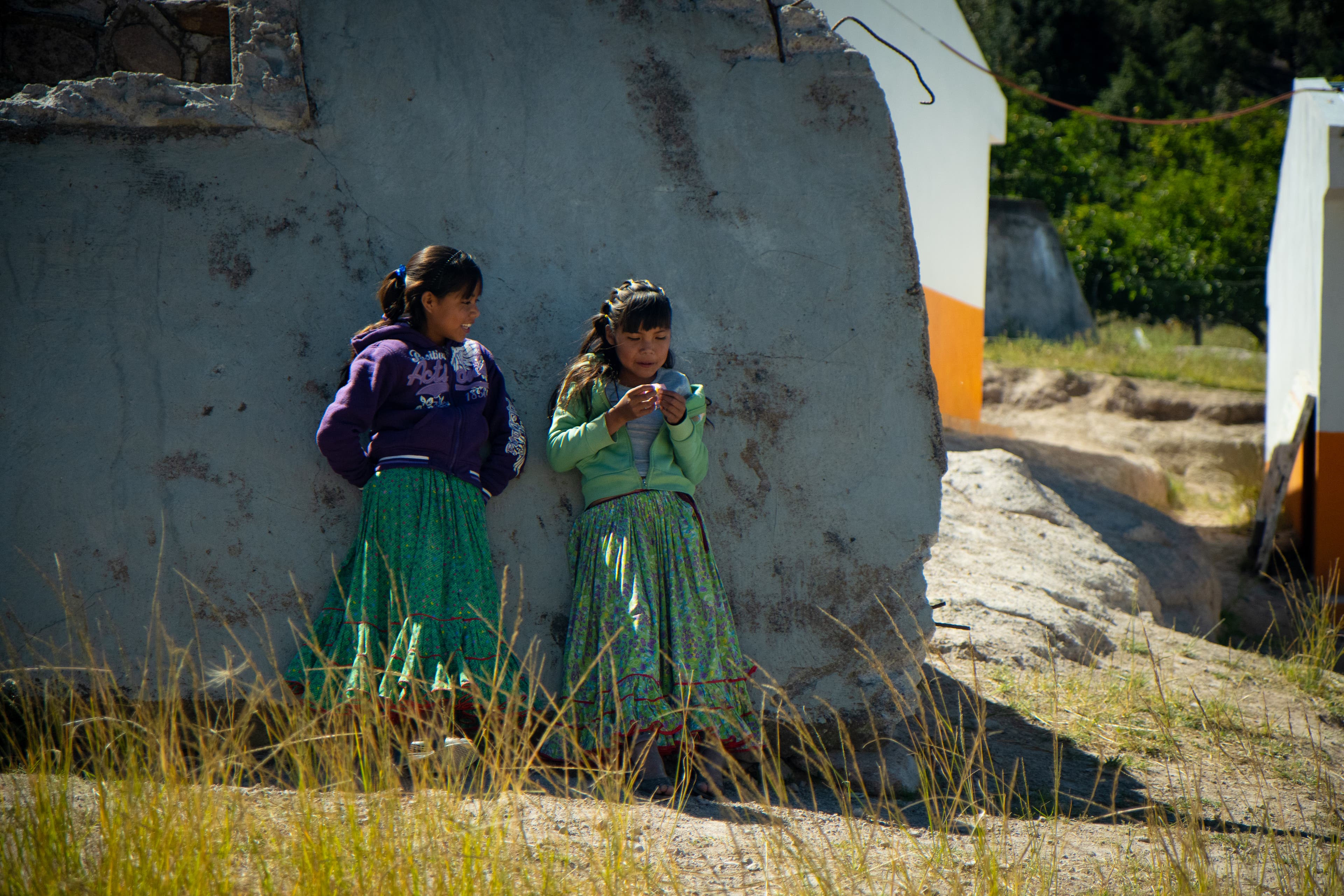 Two tween Tarahumara Indian girls in long patterned skirts lean against a concrete wall laughing with each other.