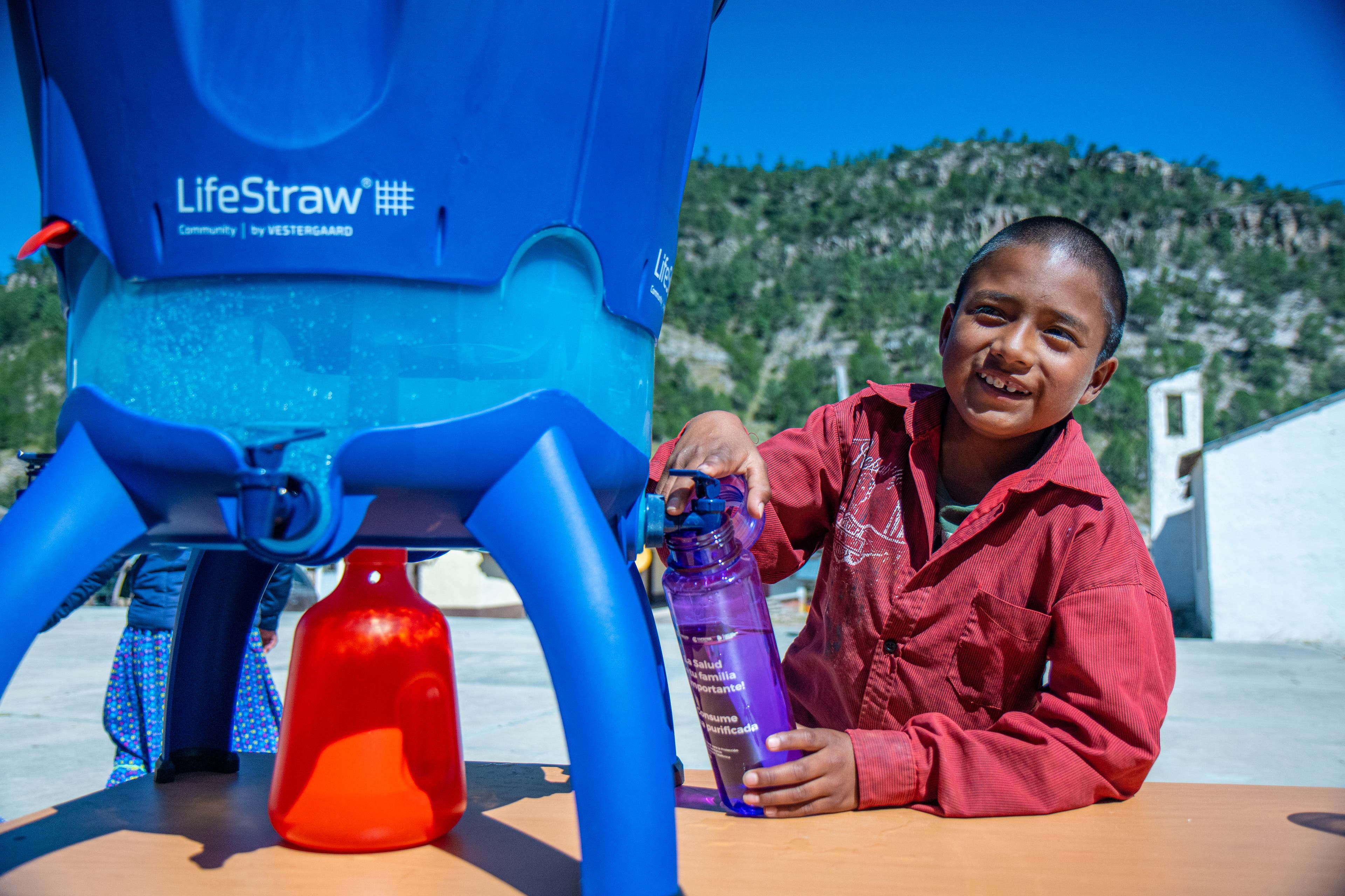 Elementary school aged Tarahumara Indian boy wears a red button down shirt and fills his water bottle from the water-cooler sized Lifestraw filter on…