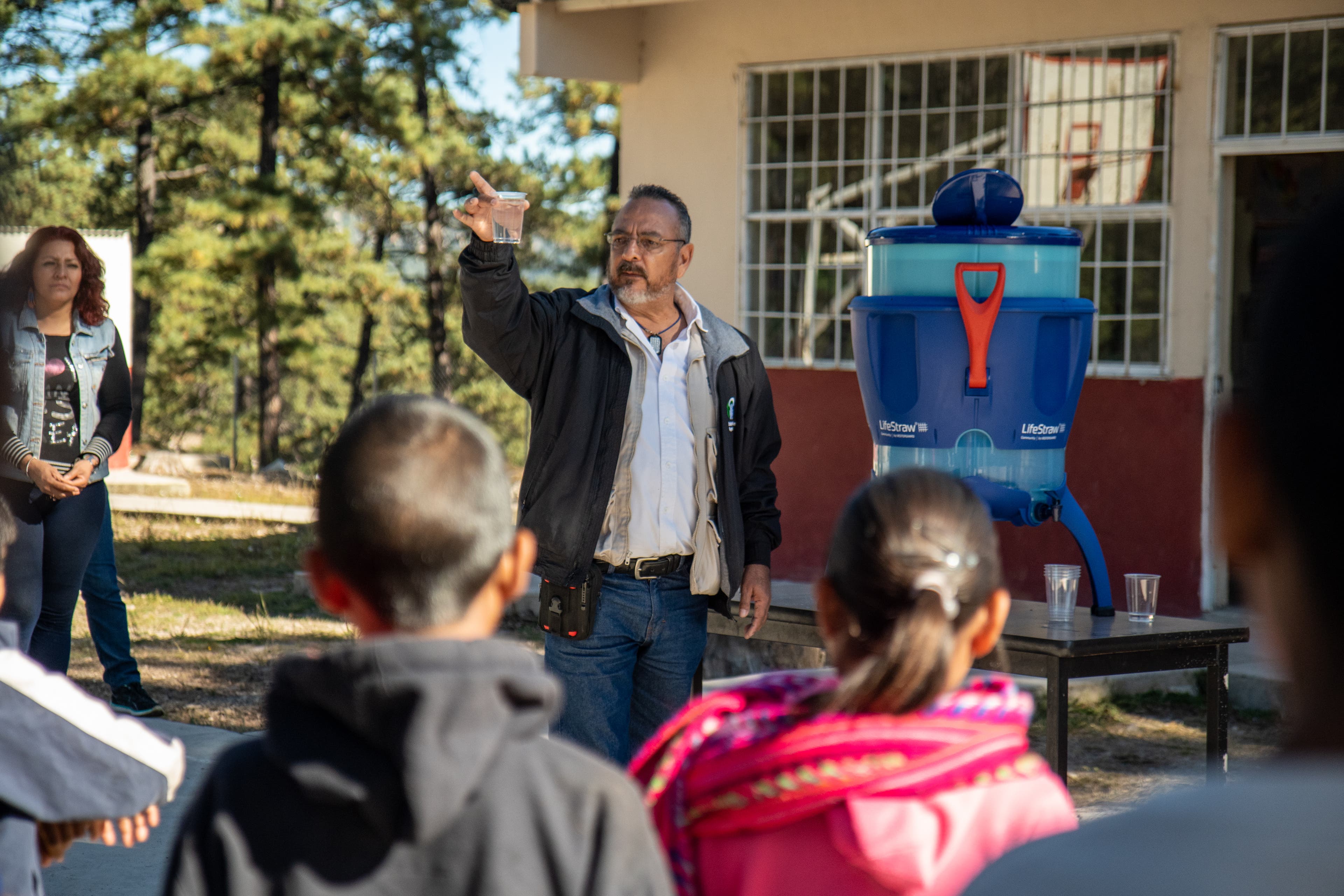 Department of Health official from Chihuahua holds up a glass of clear filtered water at the front of the presentation space in the courtyard by the…