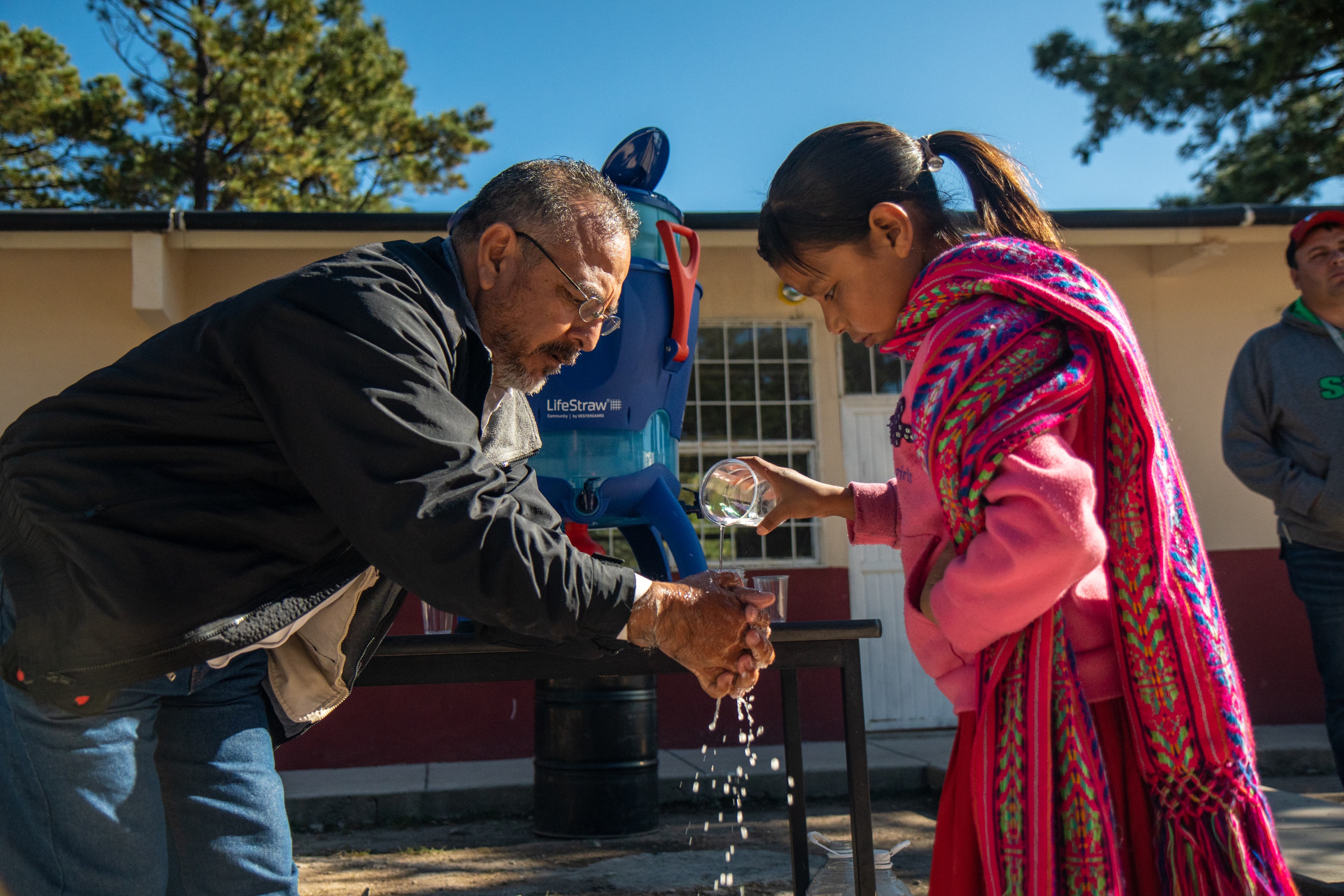 DSC09543 Man shows how to wash his hands as a young girl in a woven scarf and pink sweatshirt pours water on his hands.