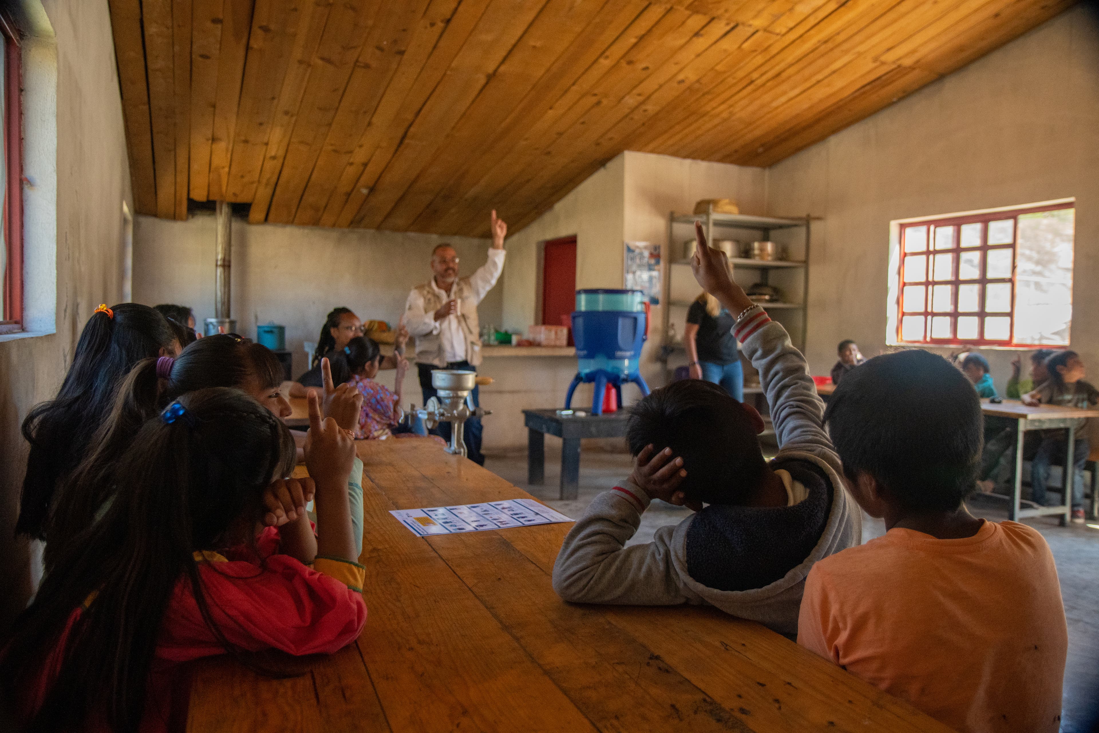Students sit at tables in a classroom while an adult man stands at the front with a cup of water in his hand. Students have their hands in the air.