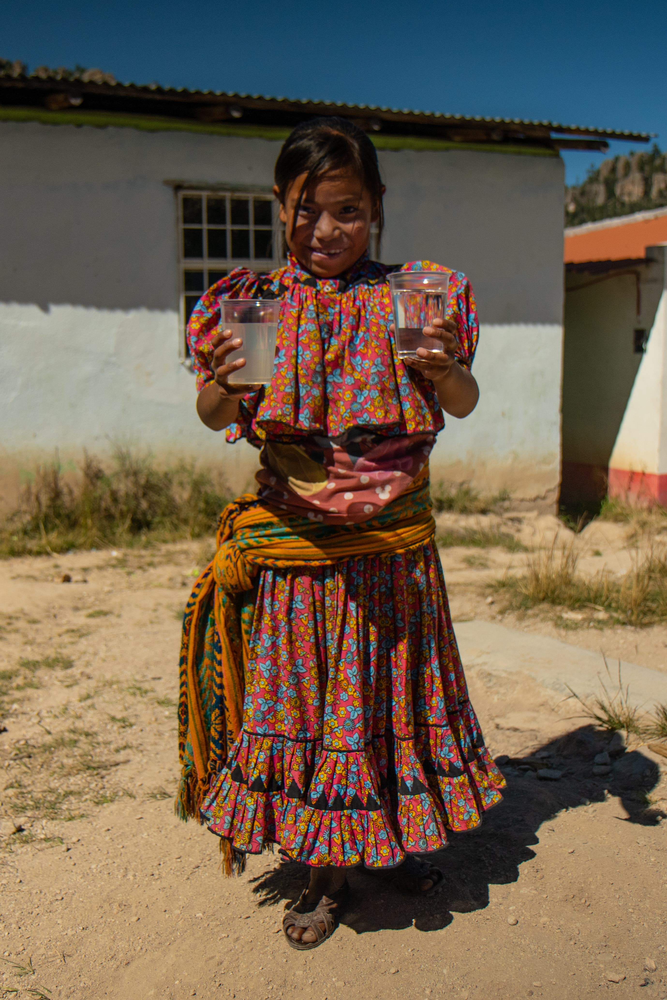 DSC09853 Ten year old Tarahumara indian girl wears colorful clothing and holds two glasses of water - one is cloudy and one is clear and filtered.