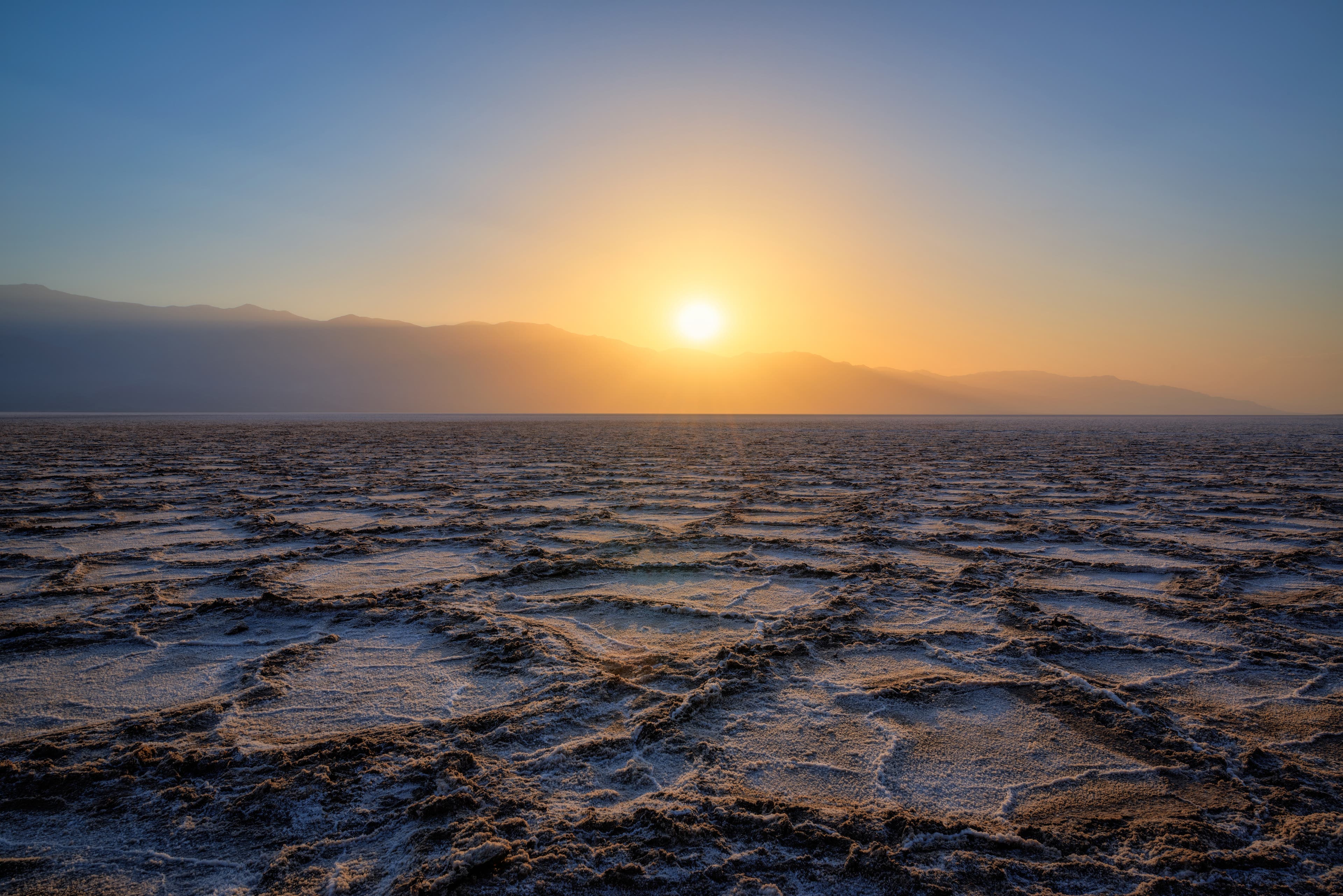 iStock-659634872_Badwater_basin The sun sets in a clear sky over the mountains in the distance at Death Valley National Park. Badwater Basin fills the foreground with its craters…