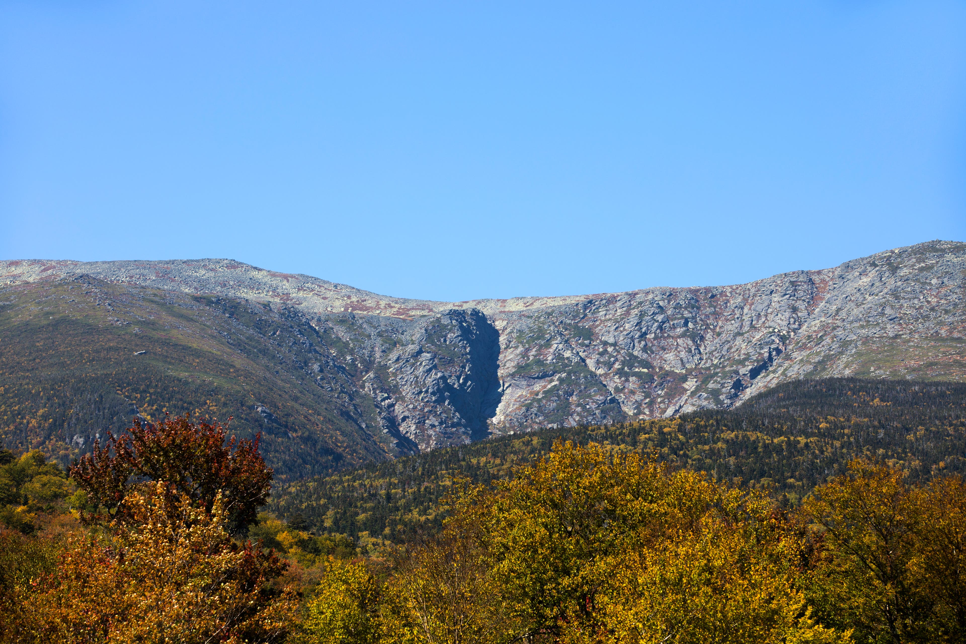 iStock-883058014_TuckermanRavine_NewHampshire Tuckerman Ravine, Mount Washington is pictured above a foreground of autumn trees. The sky is blue.