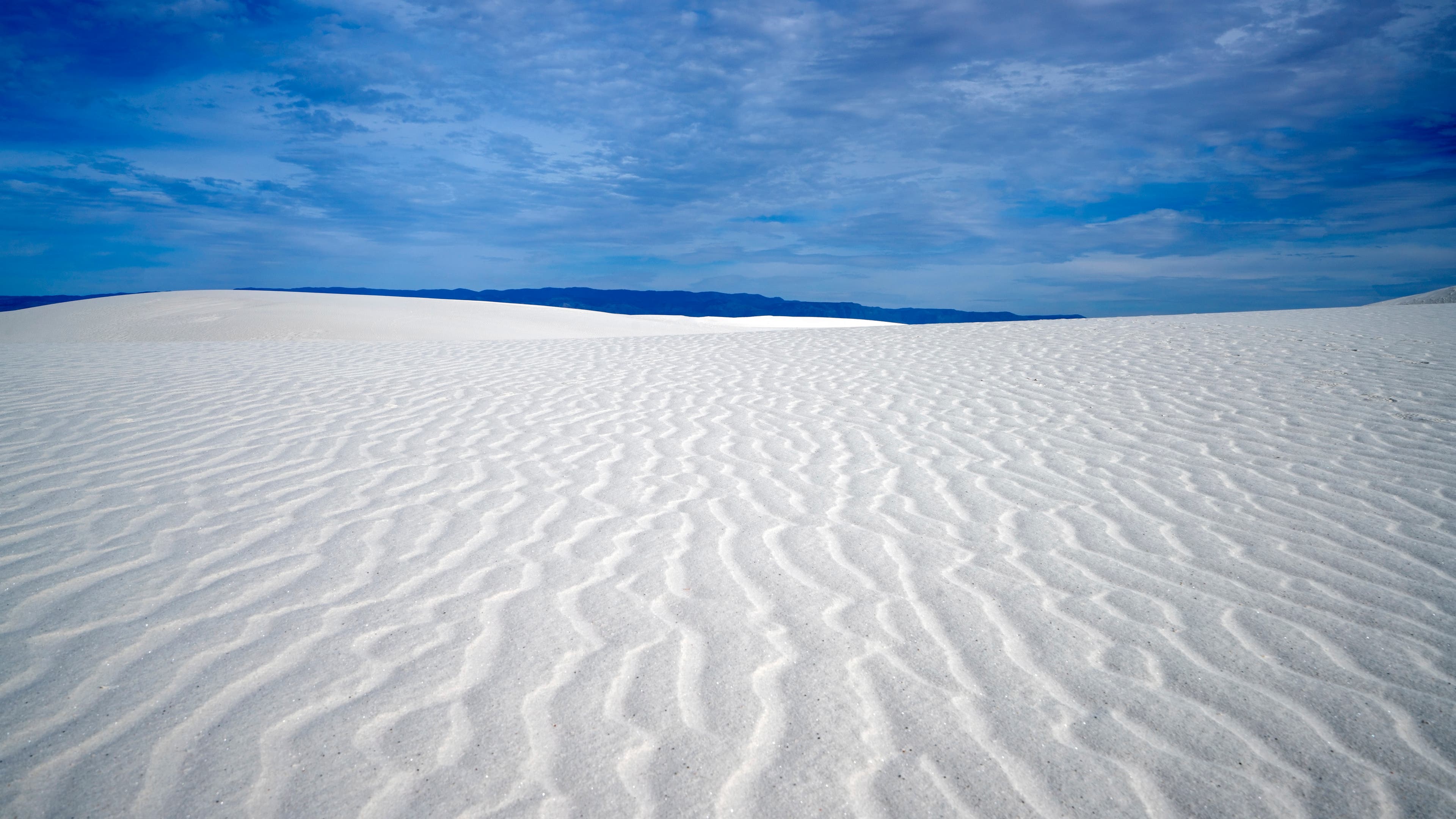 iStock-963366930_WhiteSandsMonument_AlkaliFlats Photo of white sand with ridges in it and blue skies with scattered clouds.